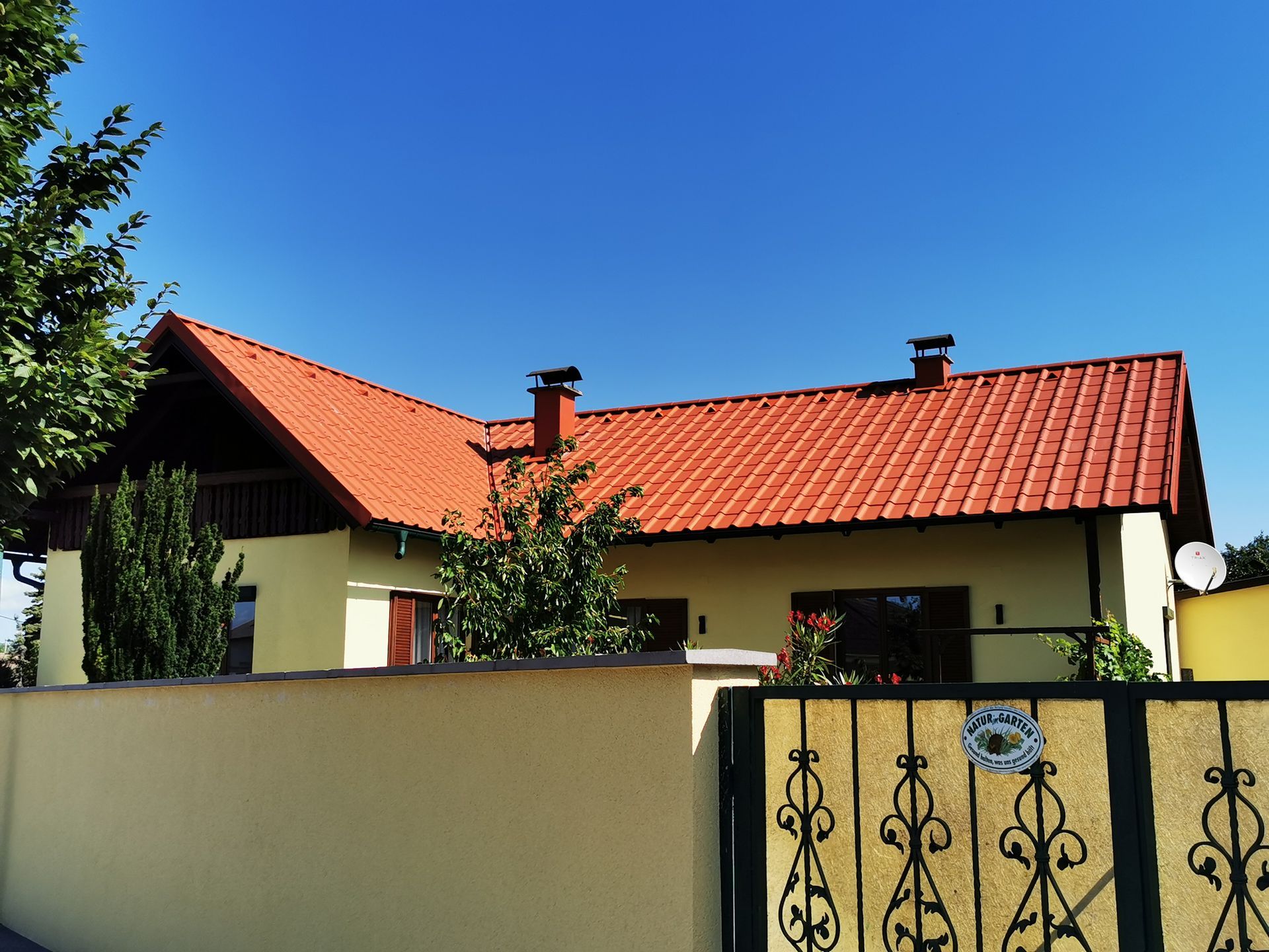 Haus mit rotem Ziegeldach hinter einer cremefarbenen Mauer und einem schmiedeeisernen Tor unter klarem, blauem Himmel