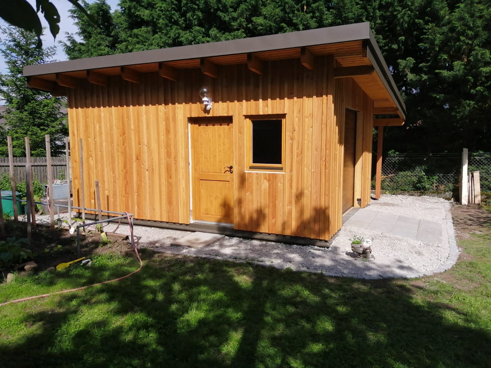 Kleiner Holzschuppen mit schrägem Dach und Fenster, in einem sonnigen Hinterhof mit Kies drumherum.
