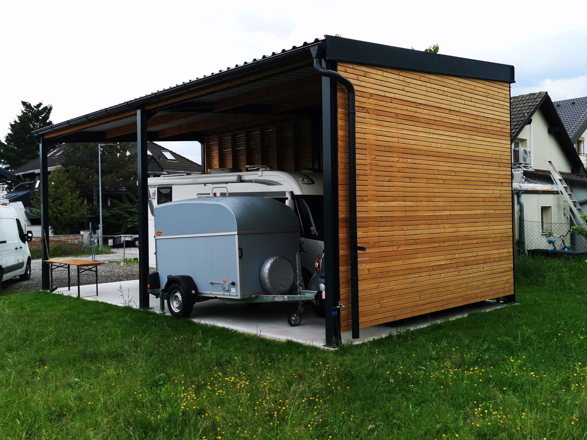 Überdachter Carport mit einem kleinen Wohnwagen und hölzernen Seitenwänden auf einem grasbewachsenen Grundstück.