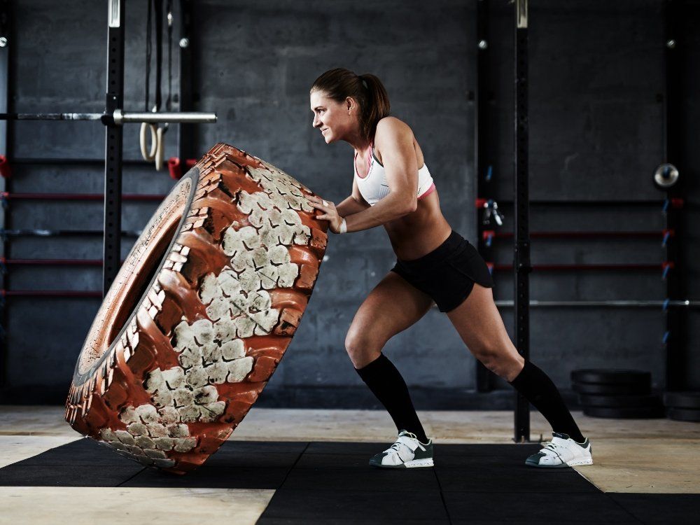 Woman in gym pushing a large tire on black floor.