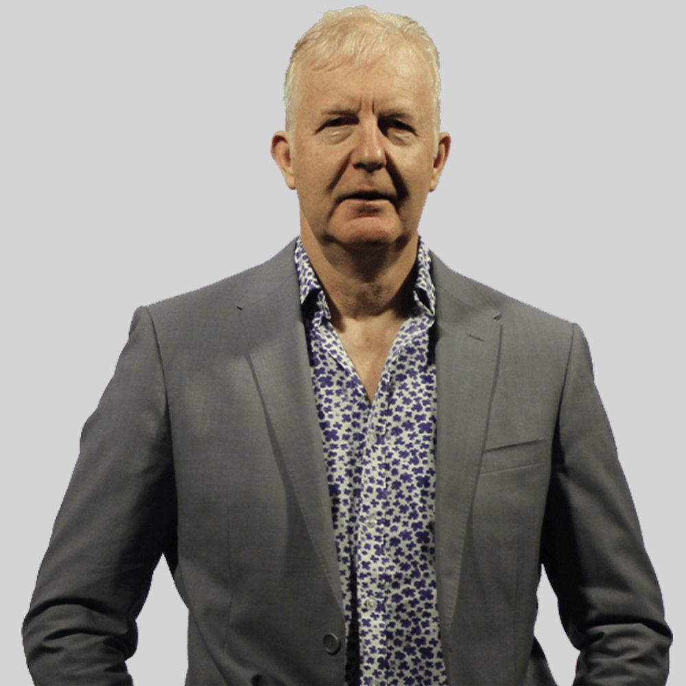 Man in gray blazer over a patterned blue and white shirt, standing against a light gray background.