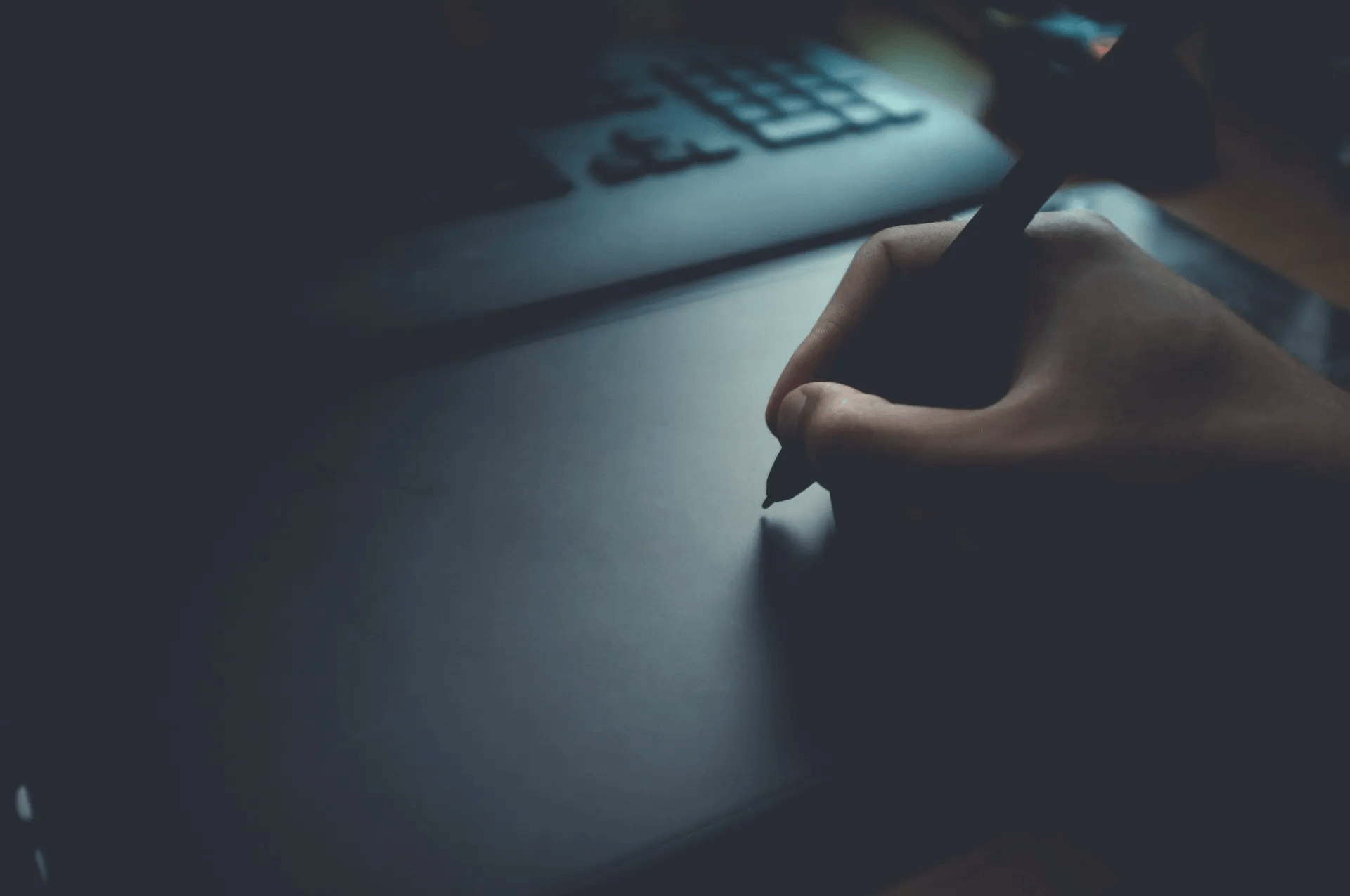 Hand using stylus on a graphic tablet, keyboard in background. Low-light setting.
