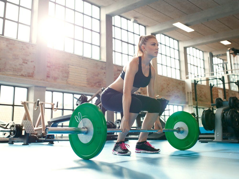Woman lifting a barbell in a gym, squatting low. Green weights on bar.