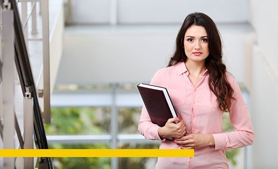 Woman in pink shirt holding a notebook, standing on a staircase, looking at the camera.