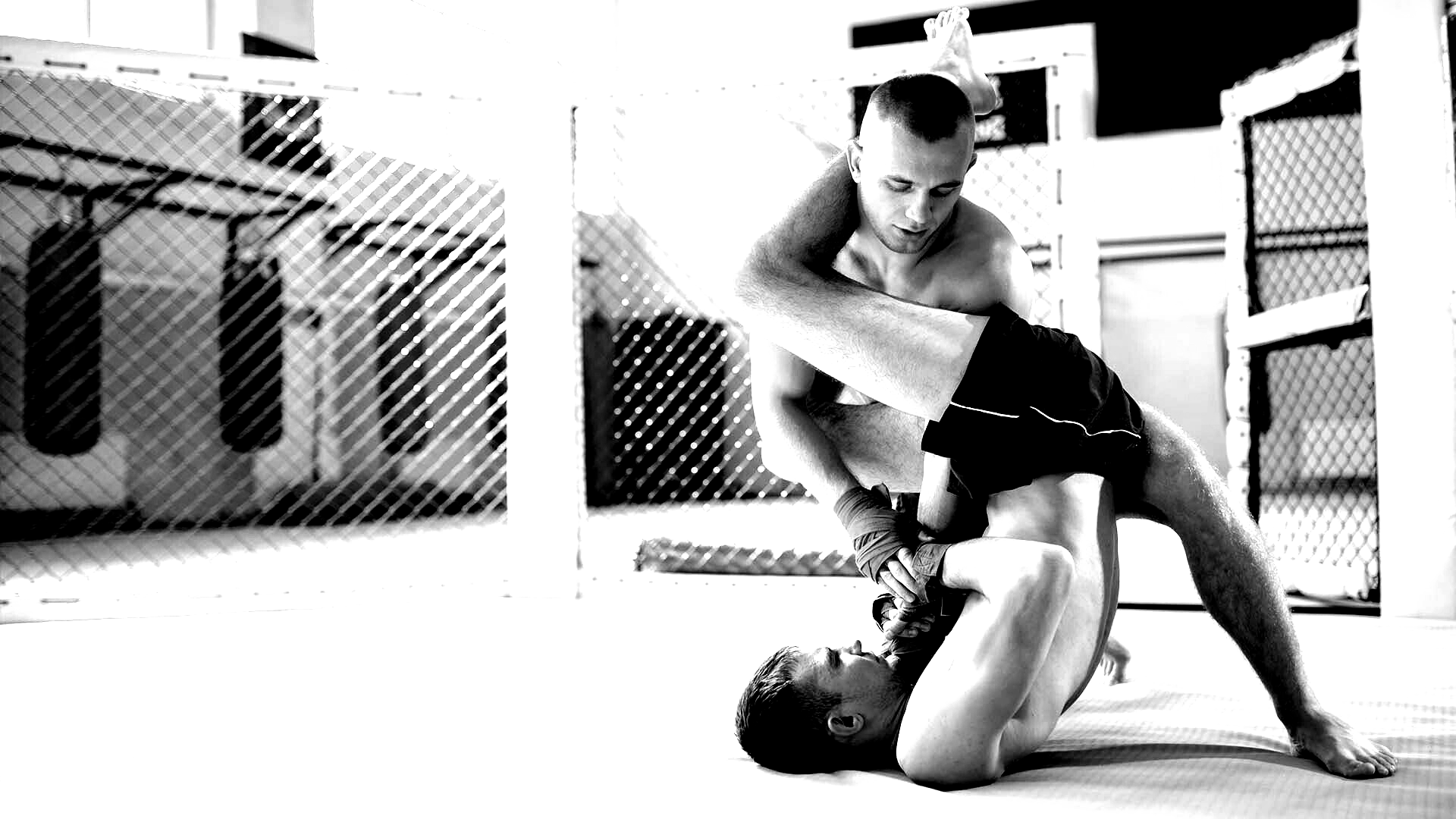 Two men grappling on a mat in a mixed martial arts cage. One has the other in a leg lock.