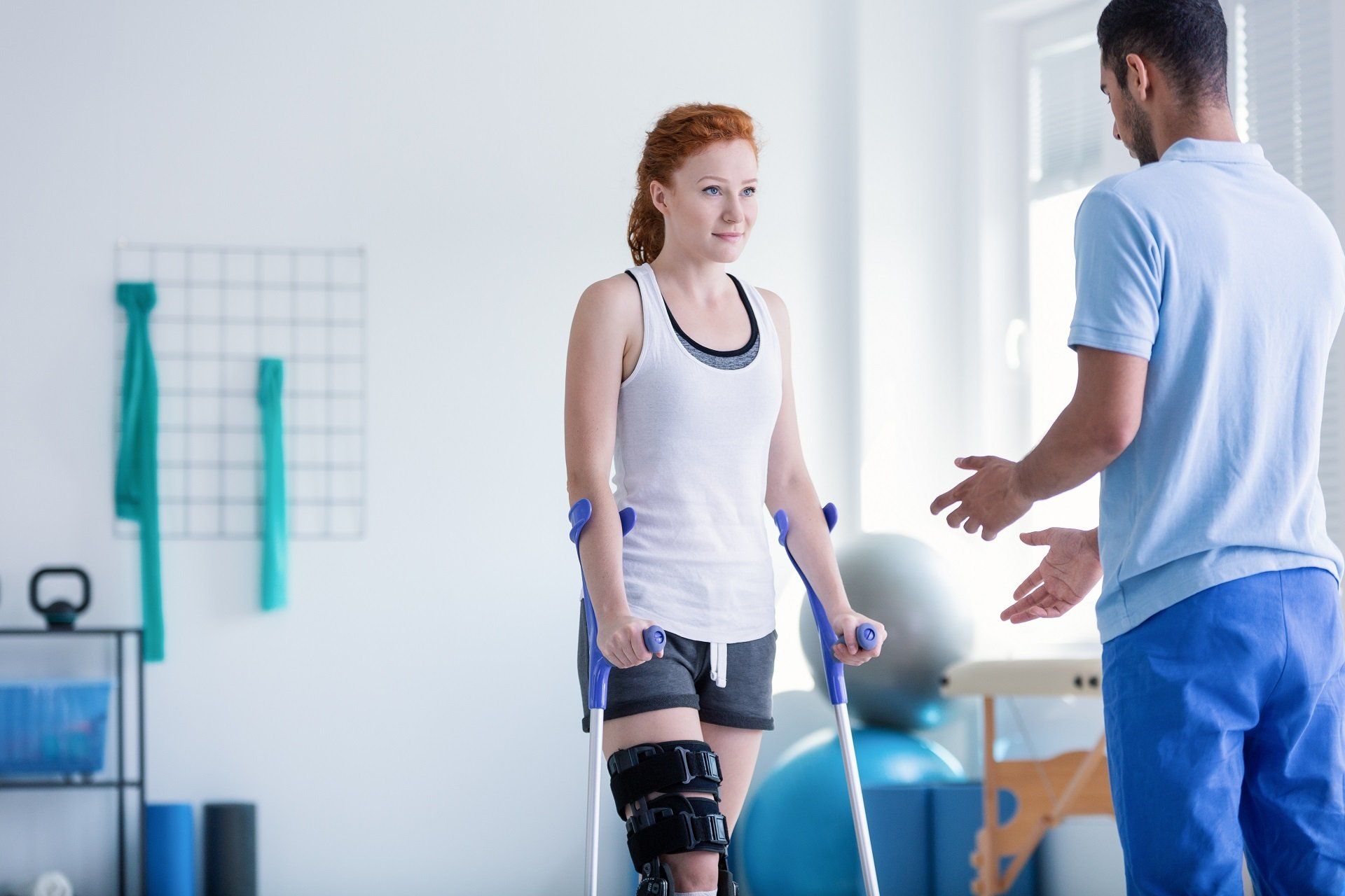 Woman with crutches and knee brace in physical therapy session, aided by a therapist in a brightly lit room.