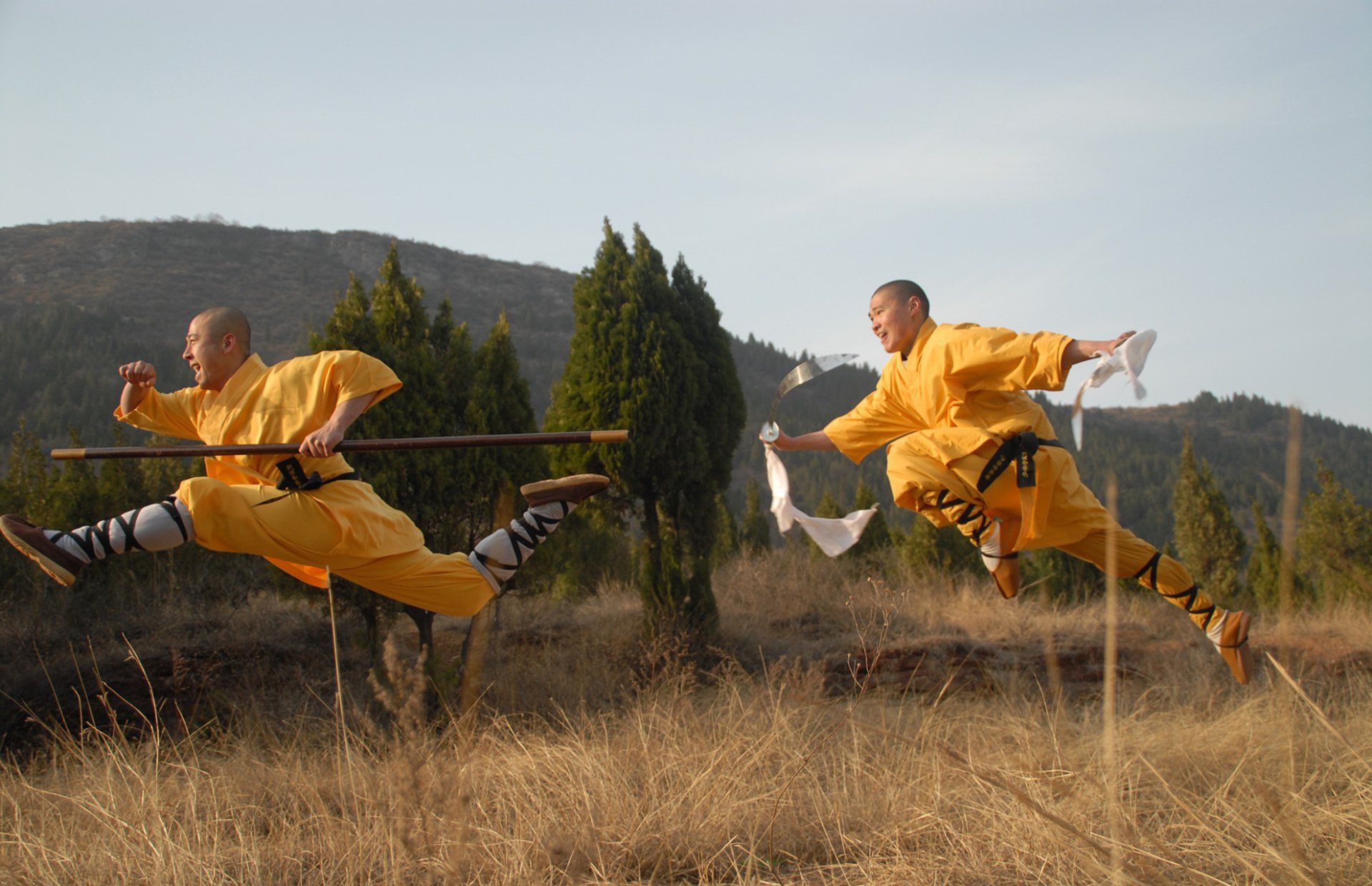 Two Shaolin monks in yellow robes leaping through dry grass. One holds a staff, the other a white cloth, against a hillside.