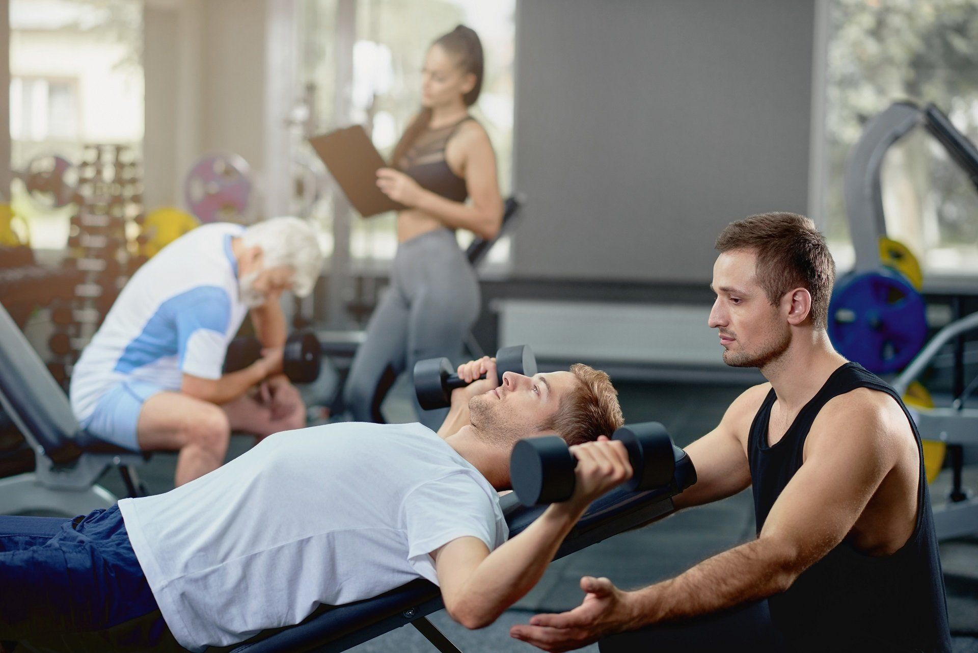 Man doing dumbbell bench press, supervised by a trainer in a gym. Another person with a clipboard.