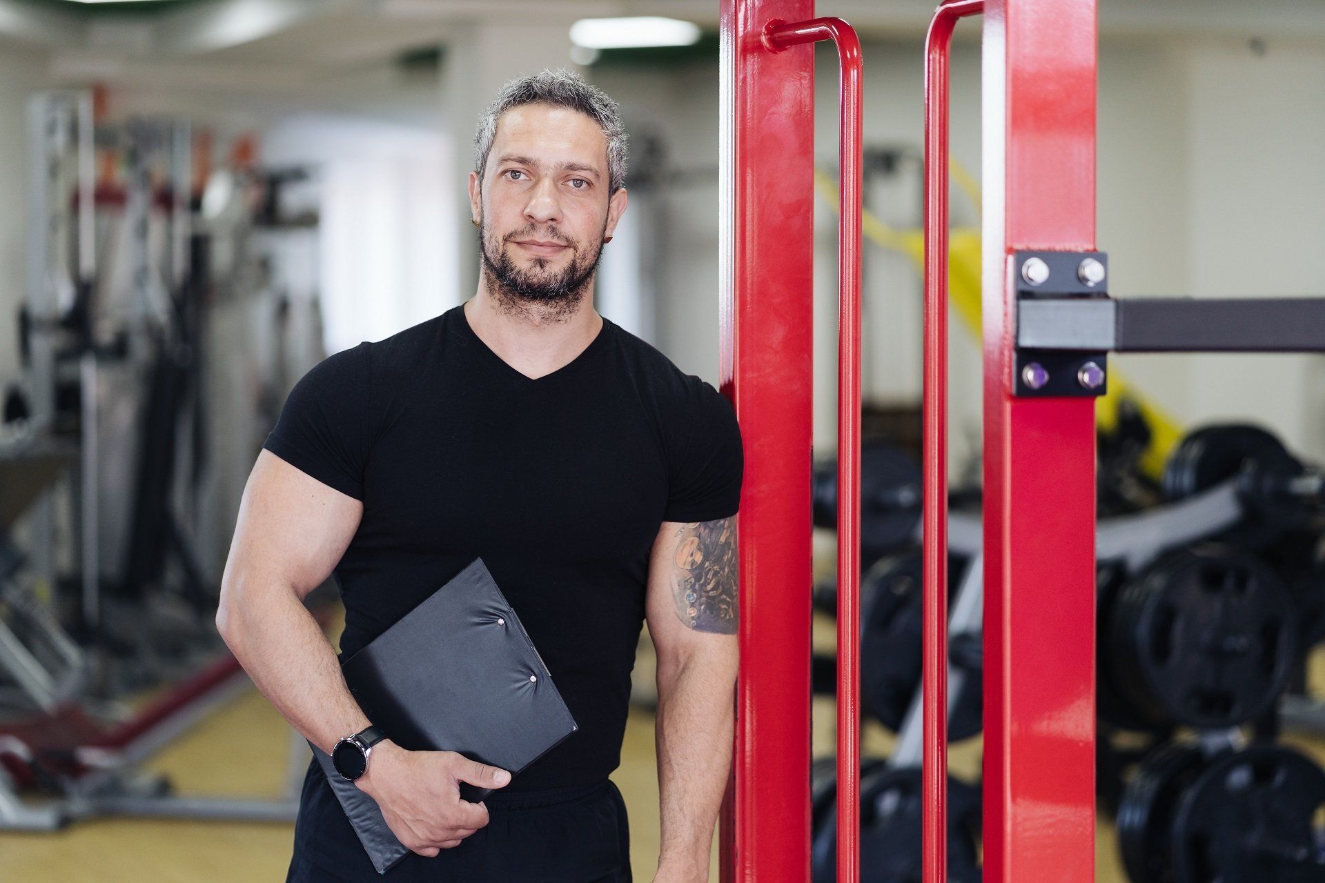 Man in black shirt with clipboard, standing by red gym equipment, smiling in a gym.