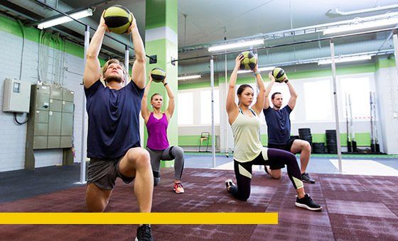People in a gym performing lunges while holding medicine balls overhead.