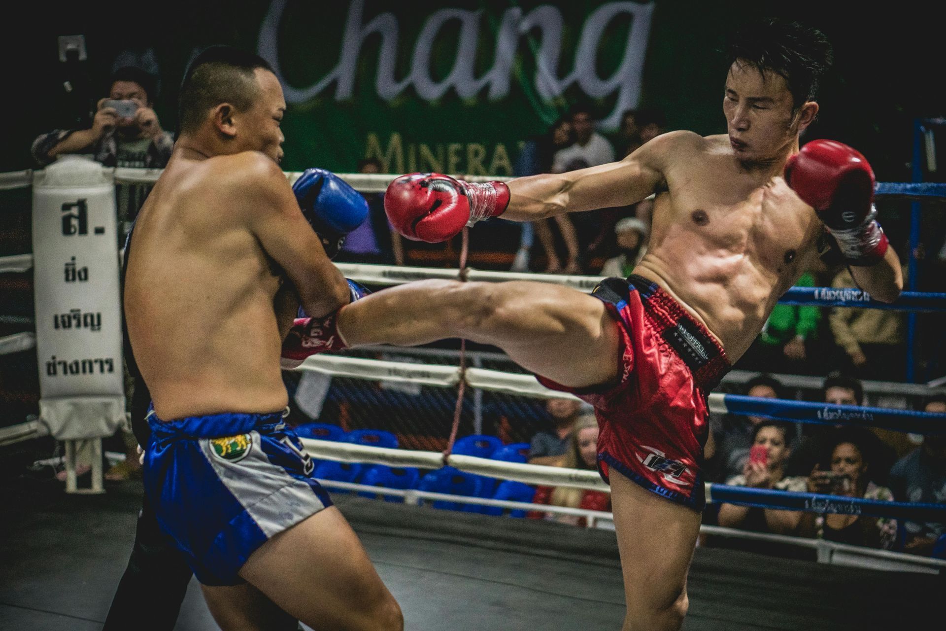 Two Muay Thai fighters spar in a ring, one kicking while the other guards in blue gloves.