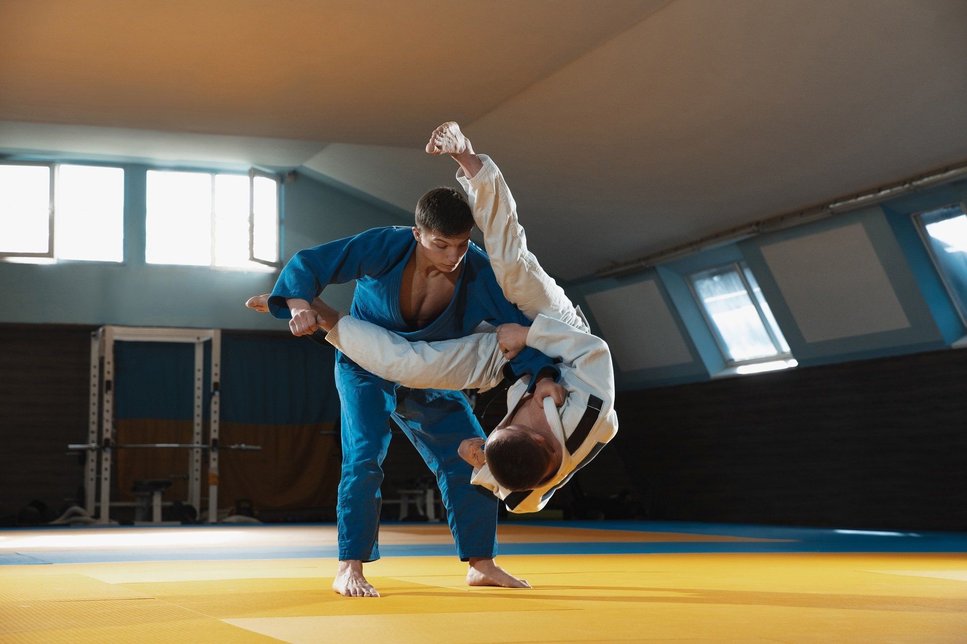 Two judo practitioners in blue and white uniforms, one throwing the other over their head on a gym mat.