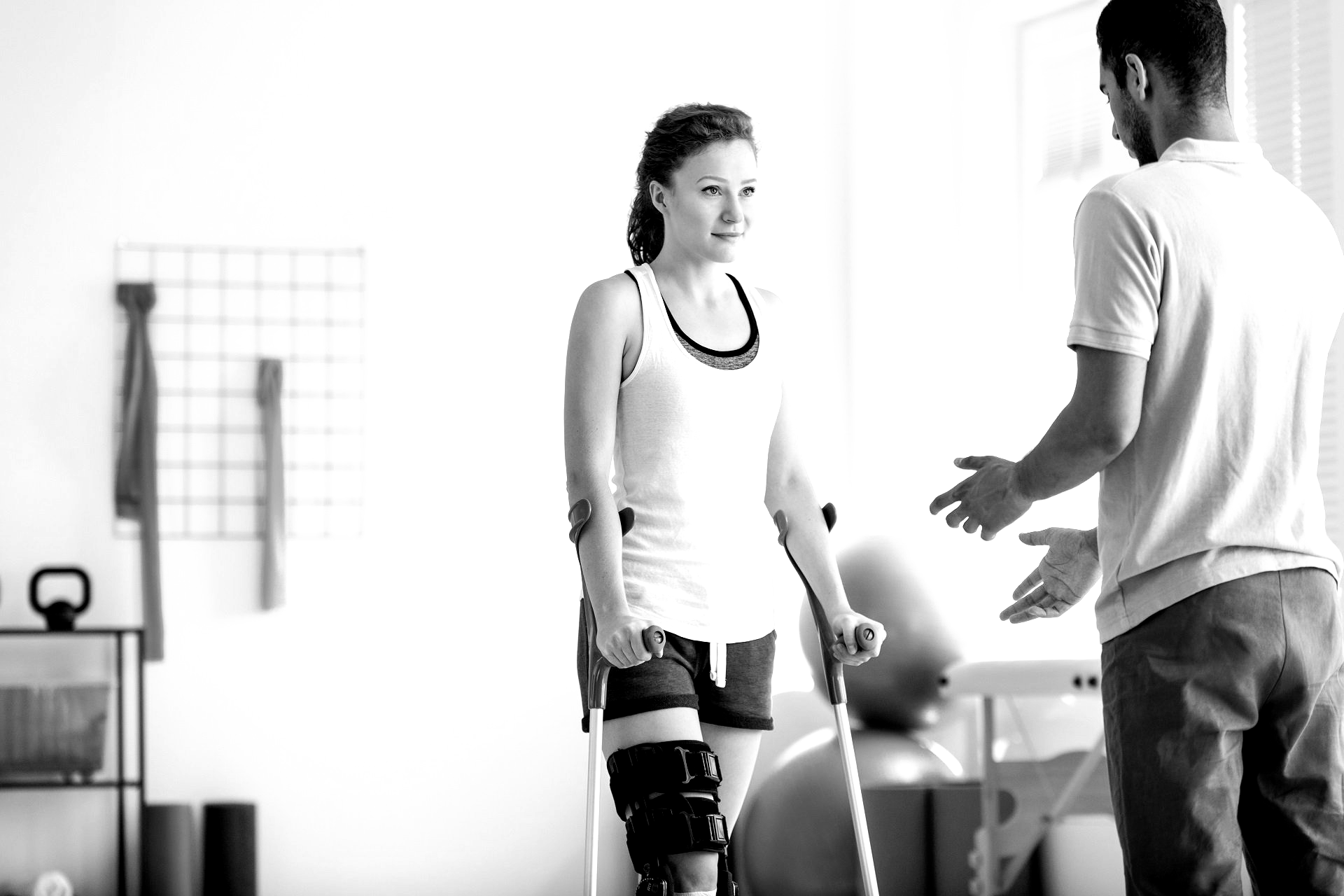 Woman with crutches and knee brace being assisted by a person in a rehabilitation setting.