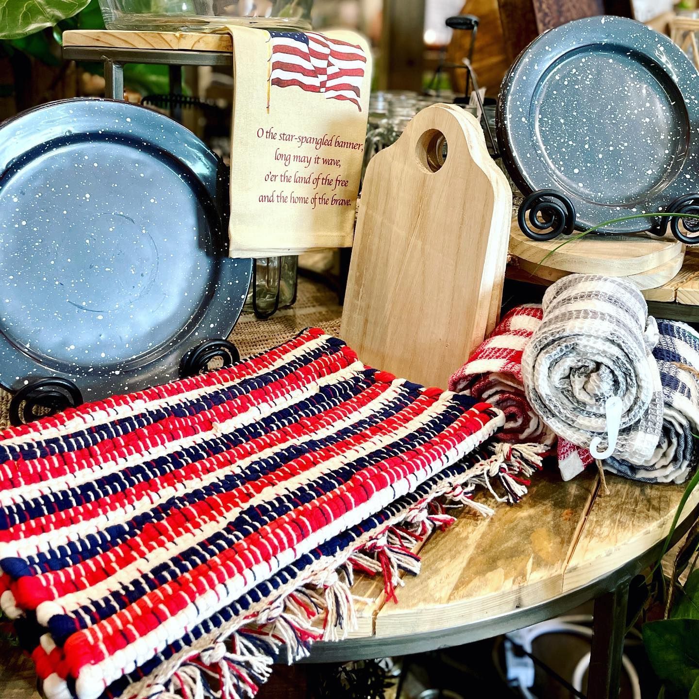 Display of red, white, and blue kitchenware, including plates, towels, cutting board, and a rug on a wooden table.