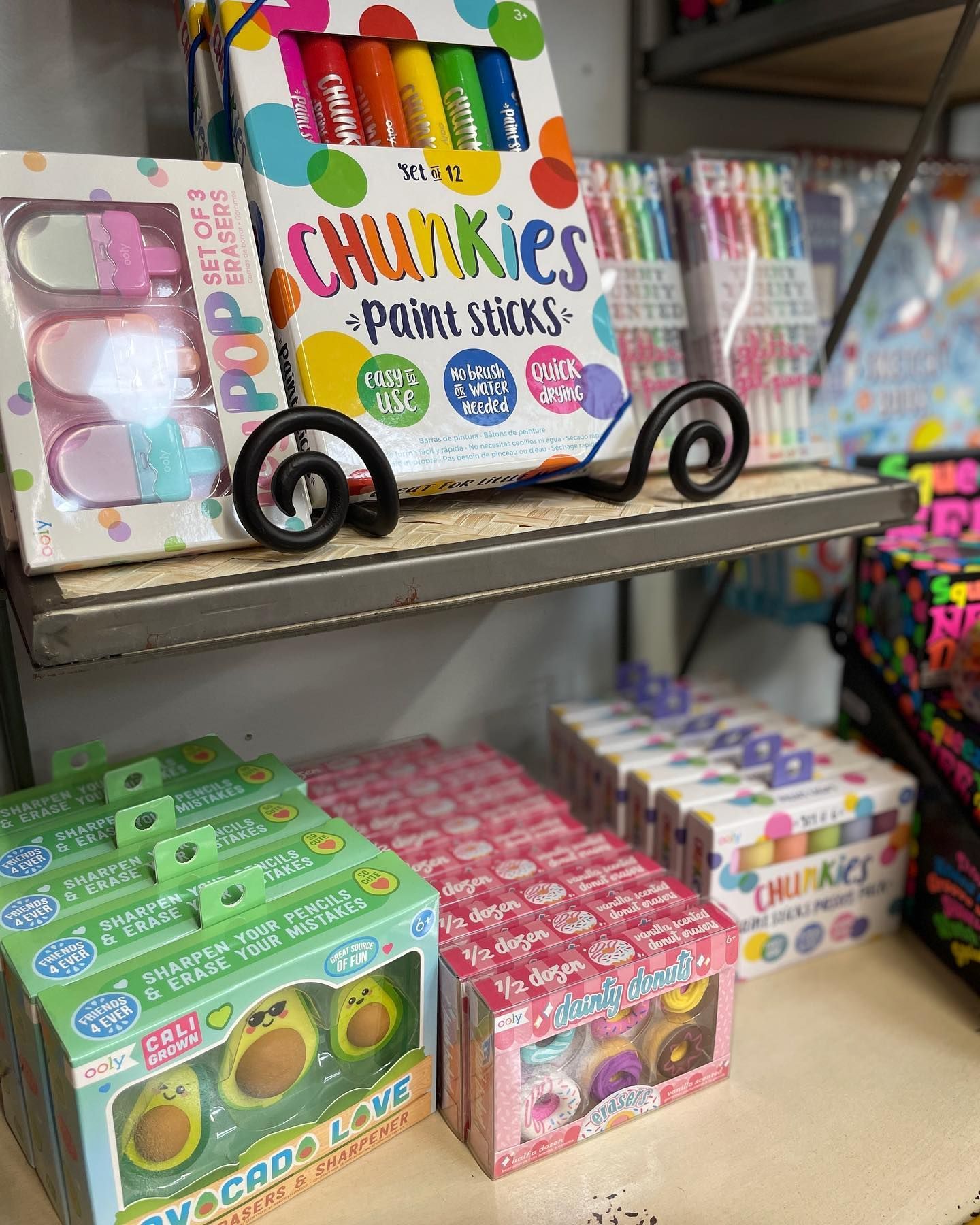 Shelf of art supplies: paint sticks, avocado erasers, donut erasers in a retail setting.