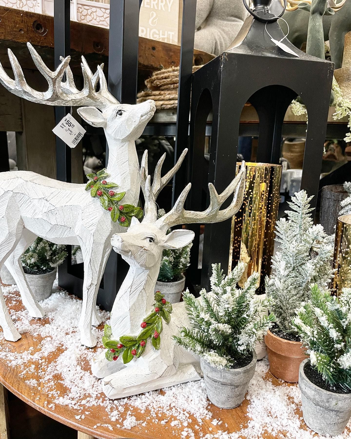 White deer statues with holiday greenery, snow, and small trees on a display table.