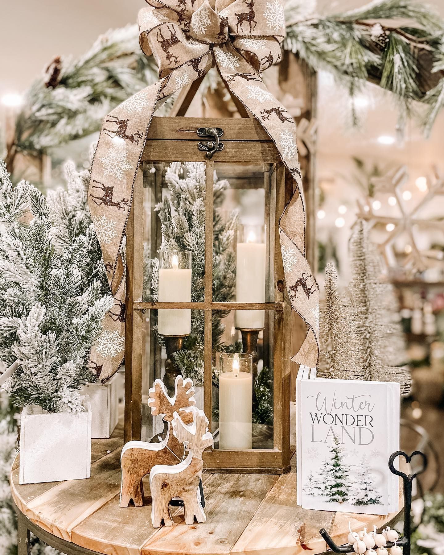 Christmas display with lit candles in a wooden lantern, snowy trees, a bow, and reindeer on a table.