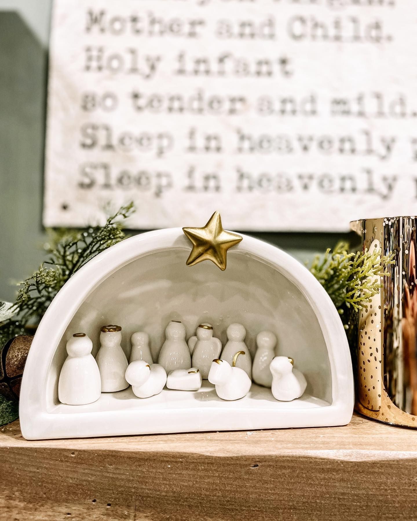 Nativity scene: white figures in a ceramic arch, topped with a gold star, with religious text in the background.