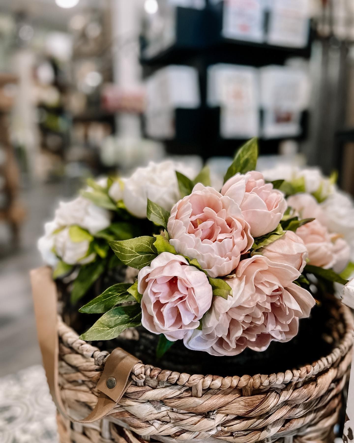 Pink and white peonies in a woven basket with a leather strap, in a store setting.