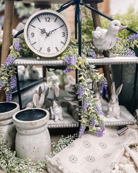 Clock, duck figurine, and bunny figurines on tiered shelf decorated with faux purple flowers and greenery.