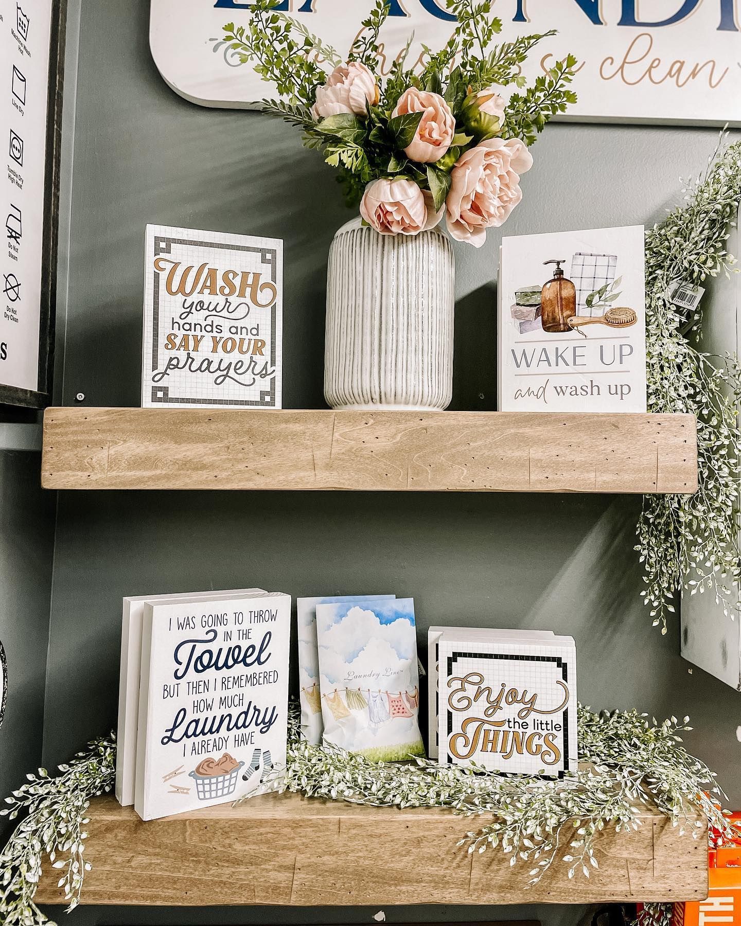 Wooden shelves with laundry-themed signs, a vase of flowers, and greenery against a gray wall.