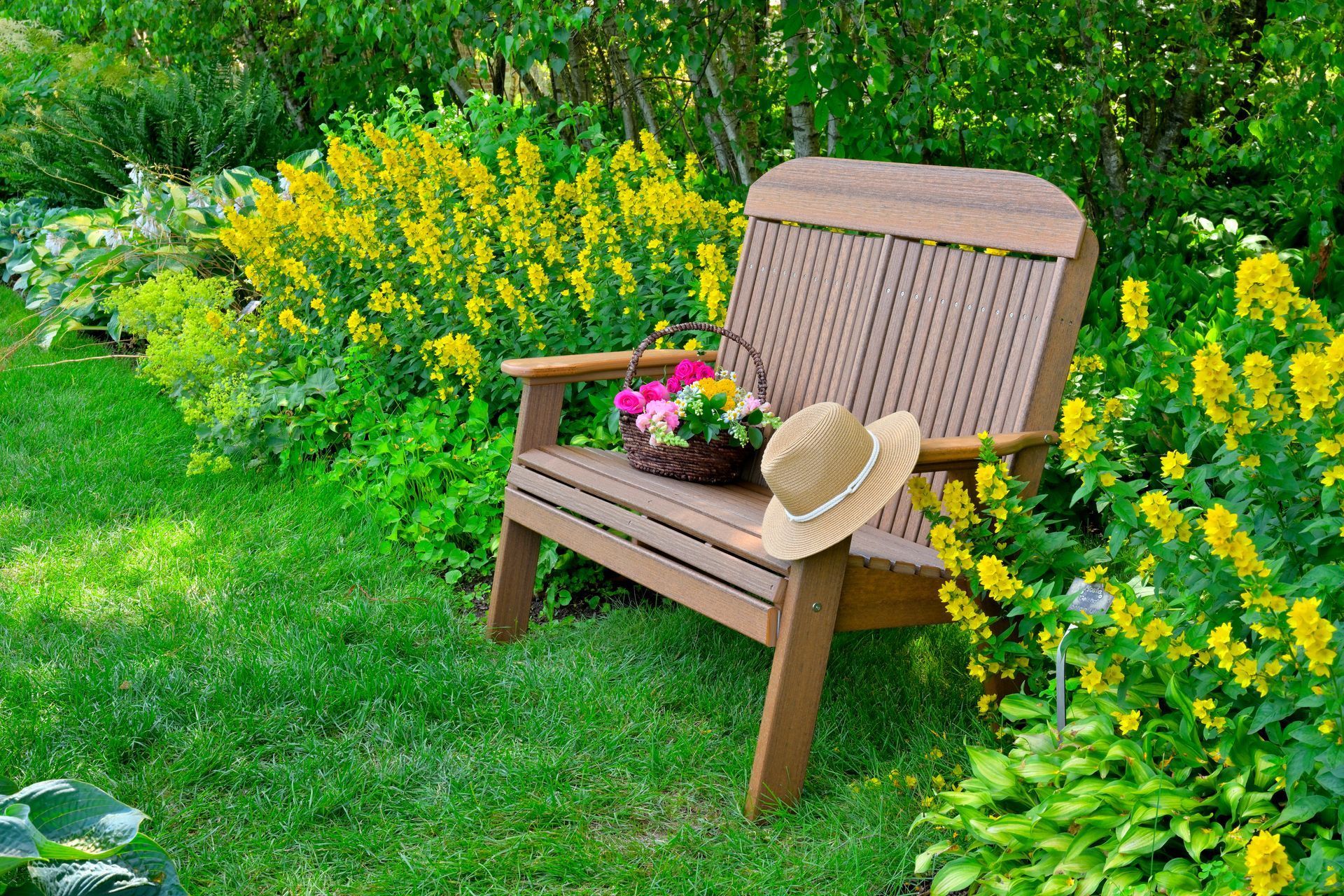 Wooden chair in a sunny garden, with basket of flowers and a straw hat. Yellow flowers and lush green foliage surround it.