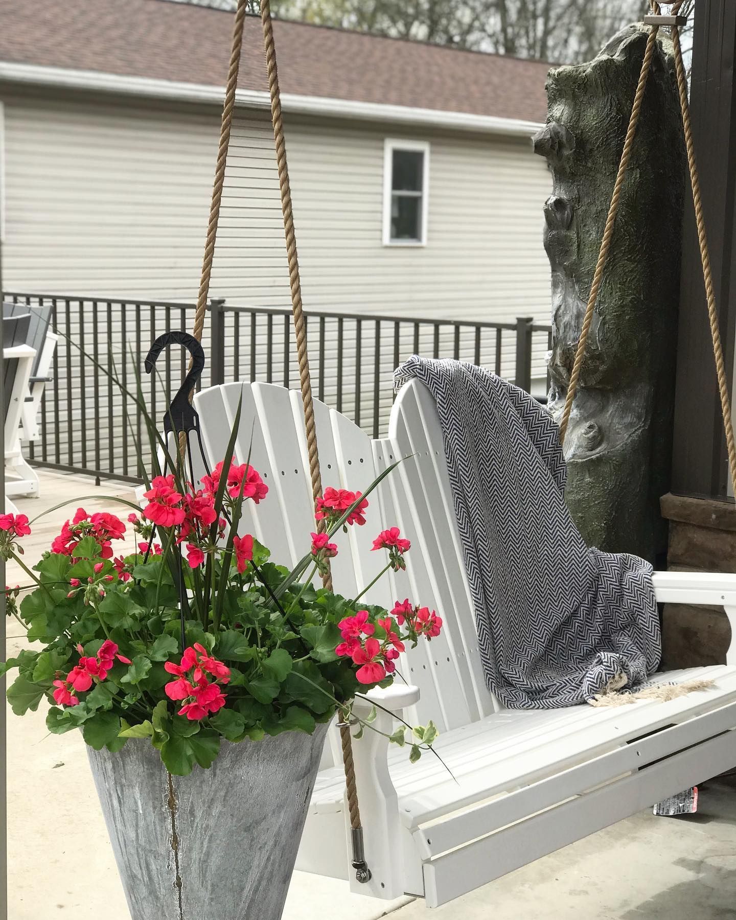 White porch swing with red geraniums, hanging from rope. Gray stone pot.