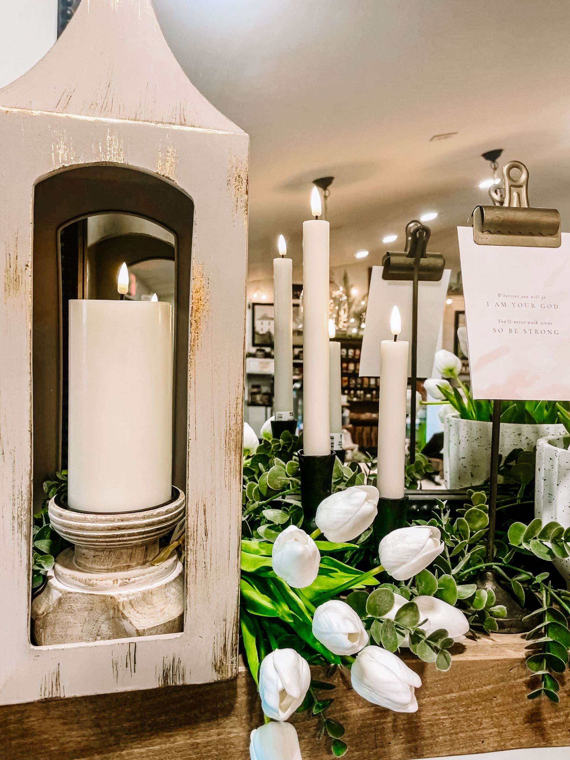 Decorative display with a lighted candle lantern, white tulips, and other candles on a table.