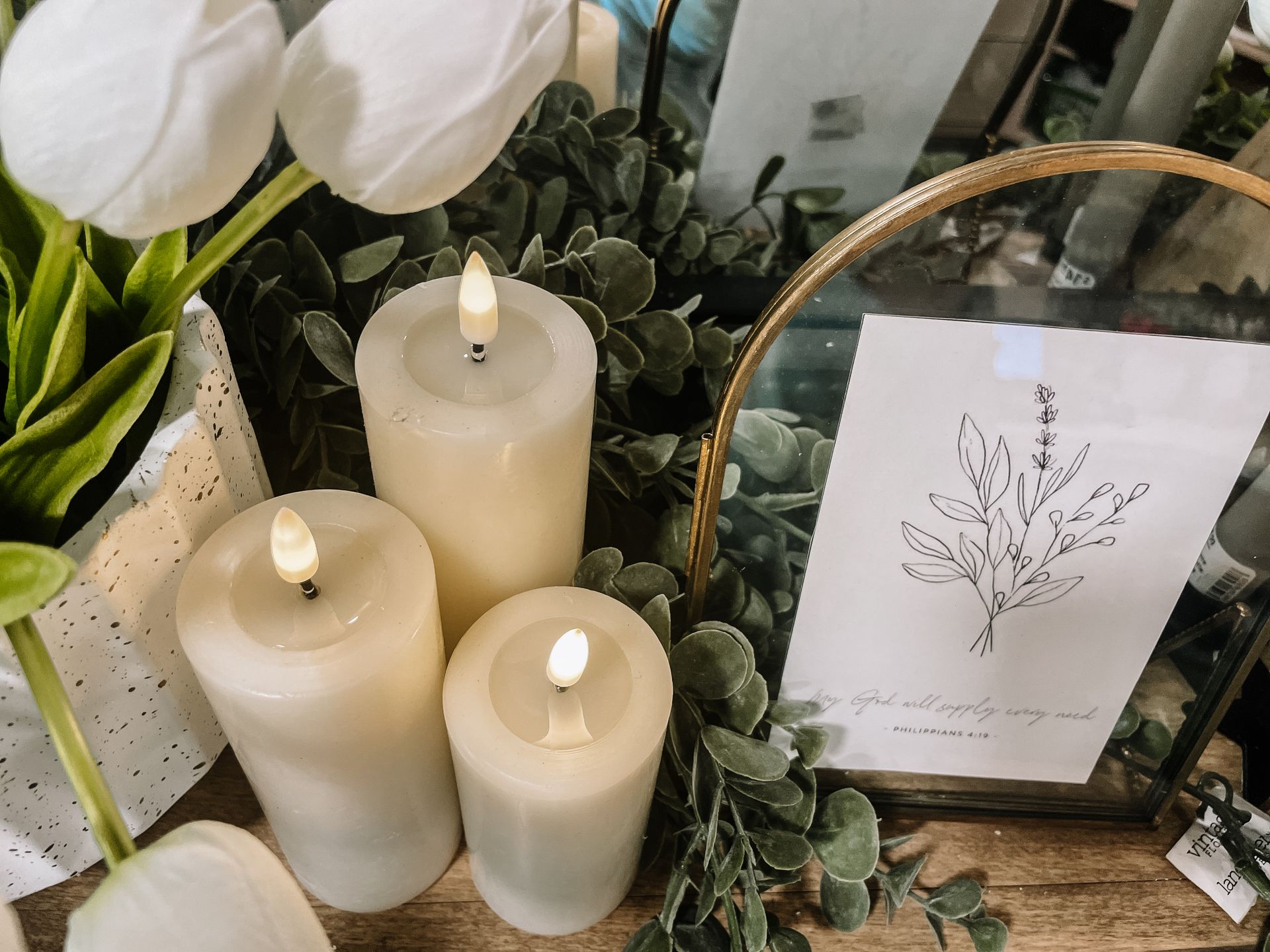 Three lit white candles, faux flowers, framed botanical print, and greenery.