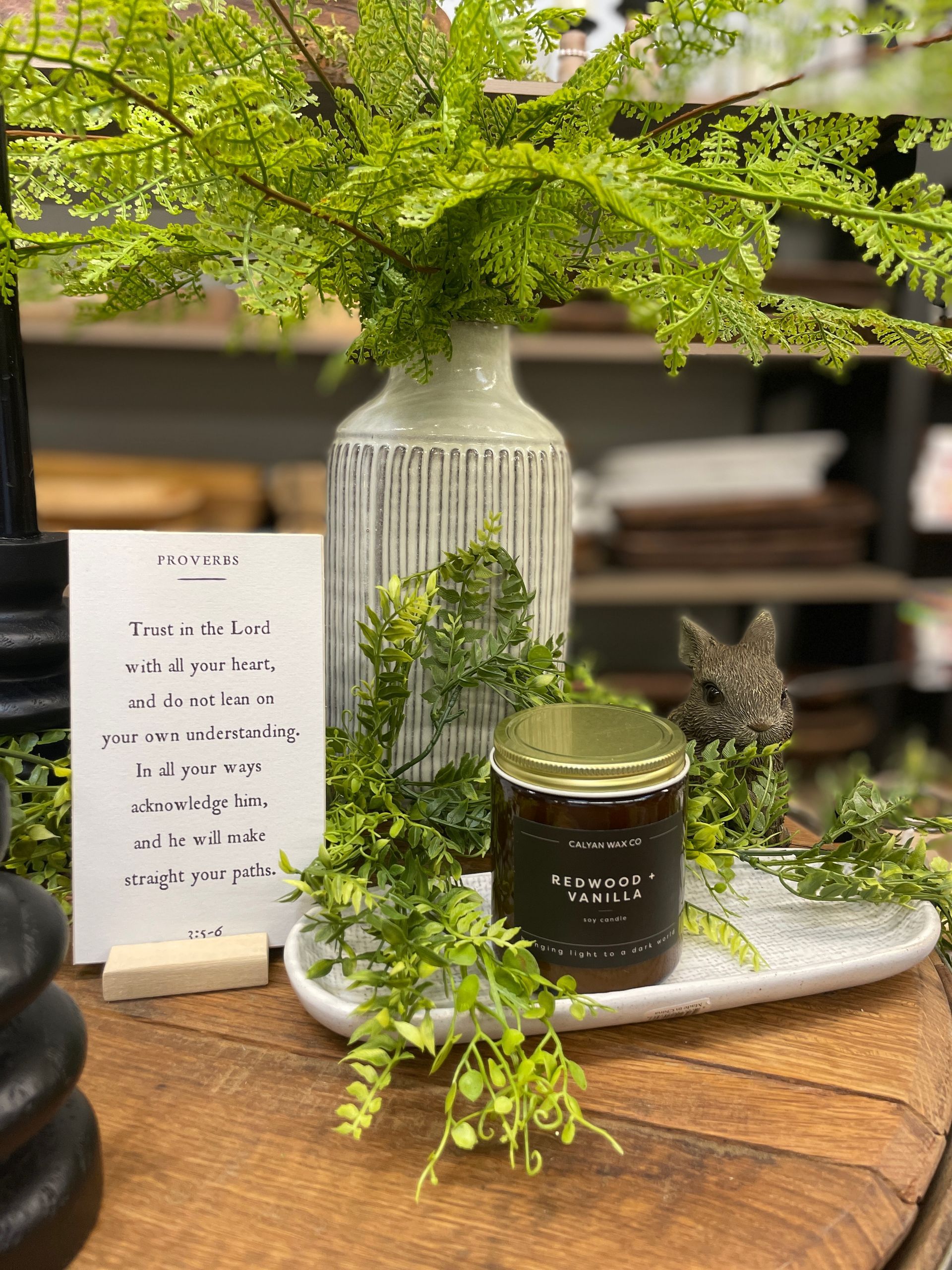 Display of decor with a vase, fern, candle, and sign on a wooden surface.