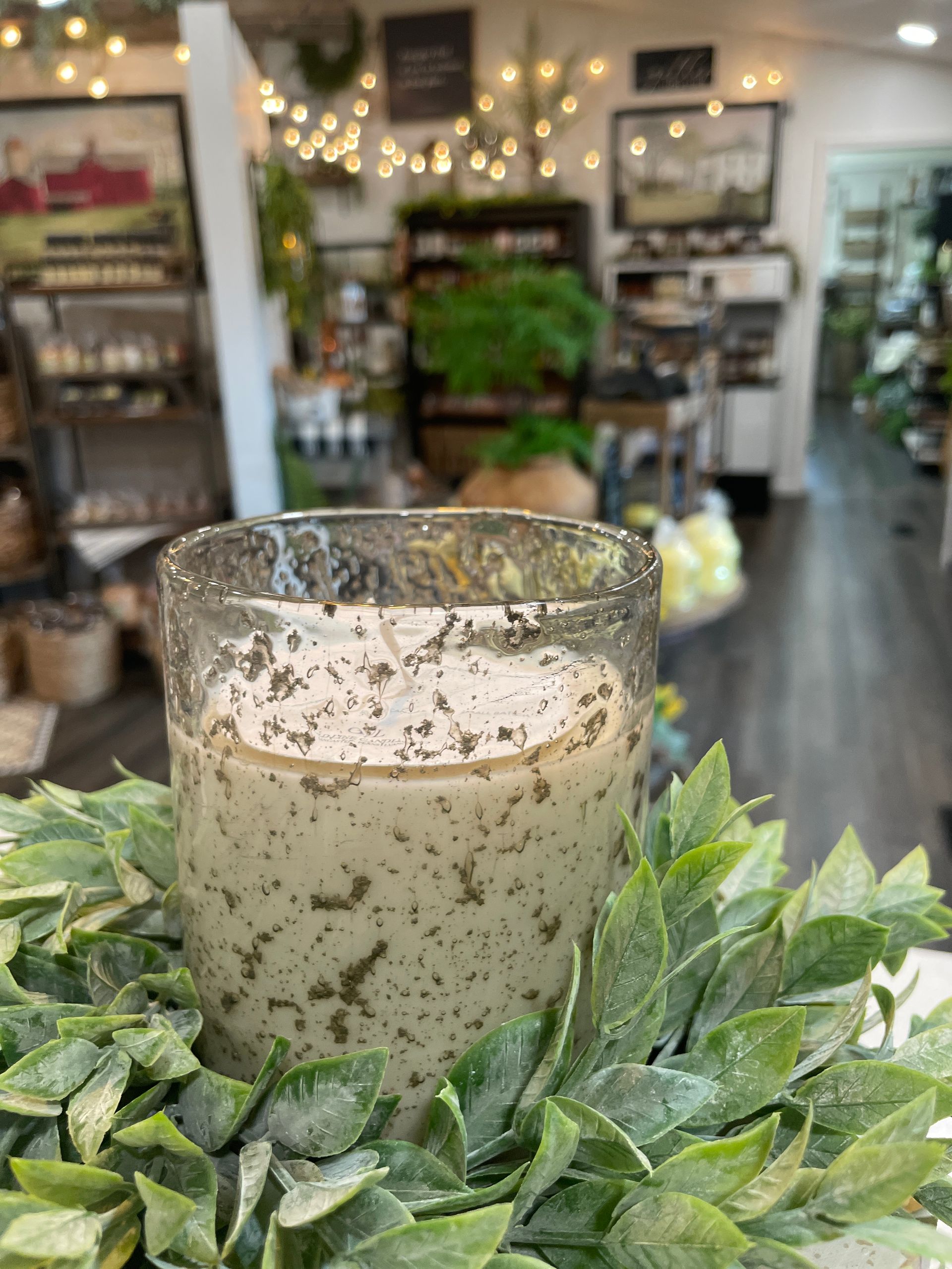 Candle speckled with green, in a glass holder, on a green leafy wreath, in a store with blurred background.