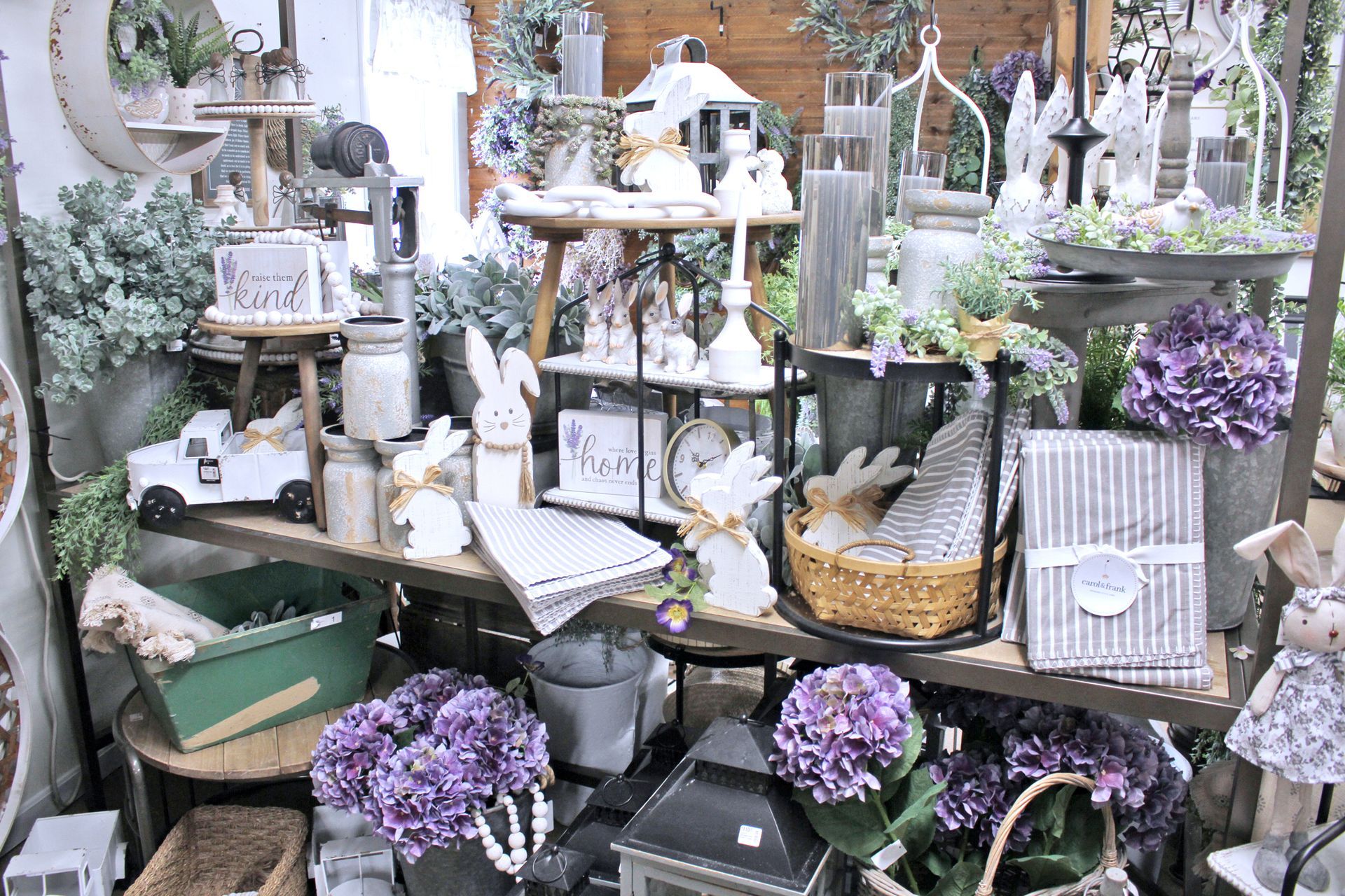 Display of Easter-themed home decor with rabbits, purple flowers, and candles on shelves and tables.