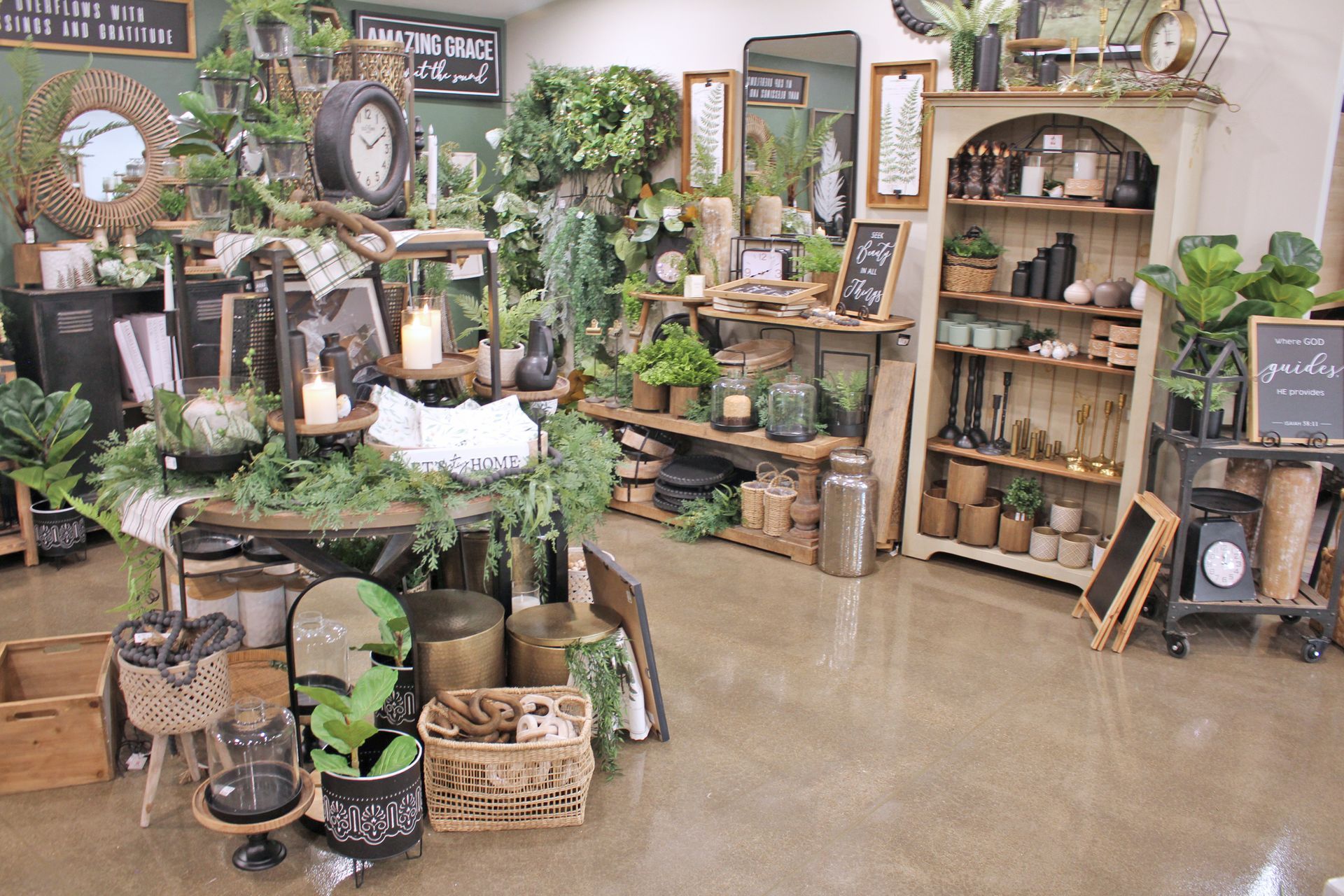 A well-lit shop interior displays home decor items, including plants, clocks, and shelves against a green wall.