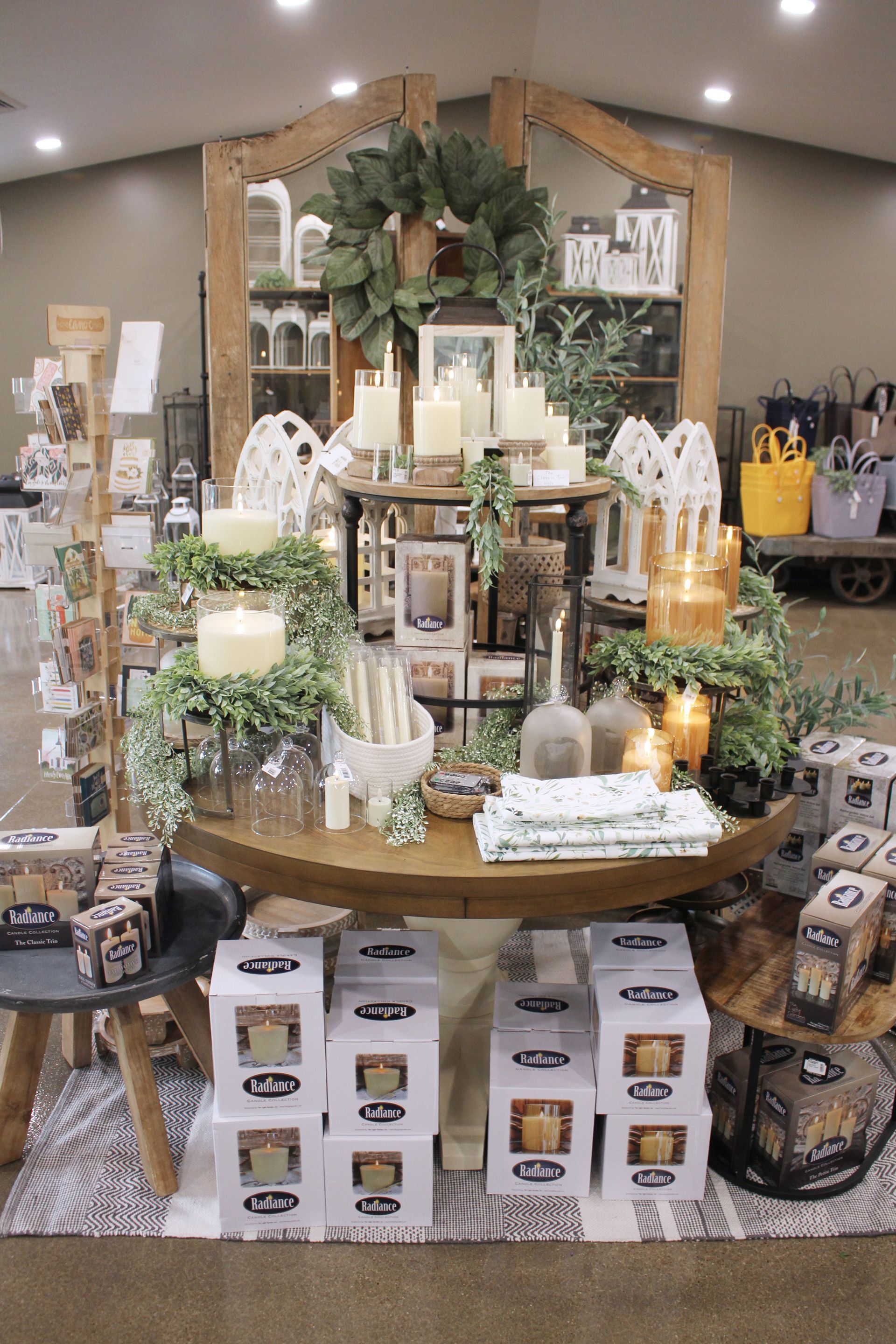 Retail display of candles and decor, with a wooden arched backdrop and greenery.