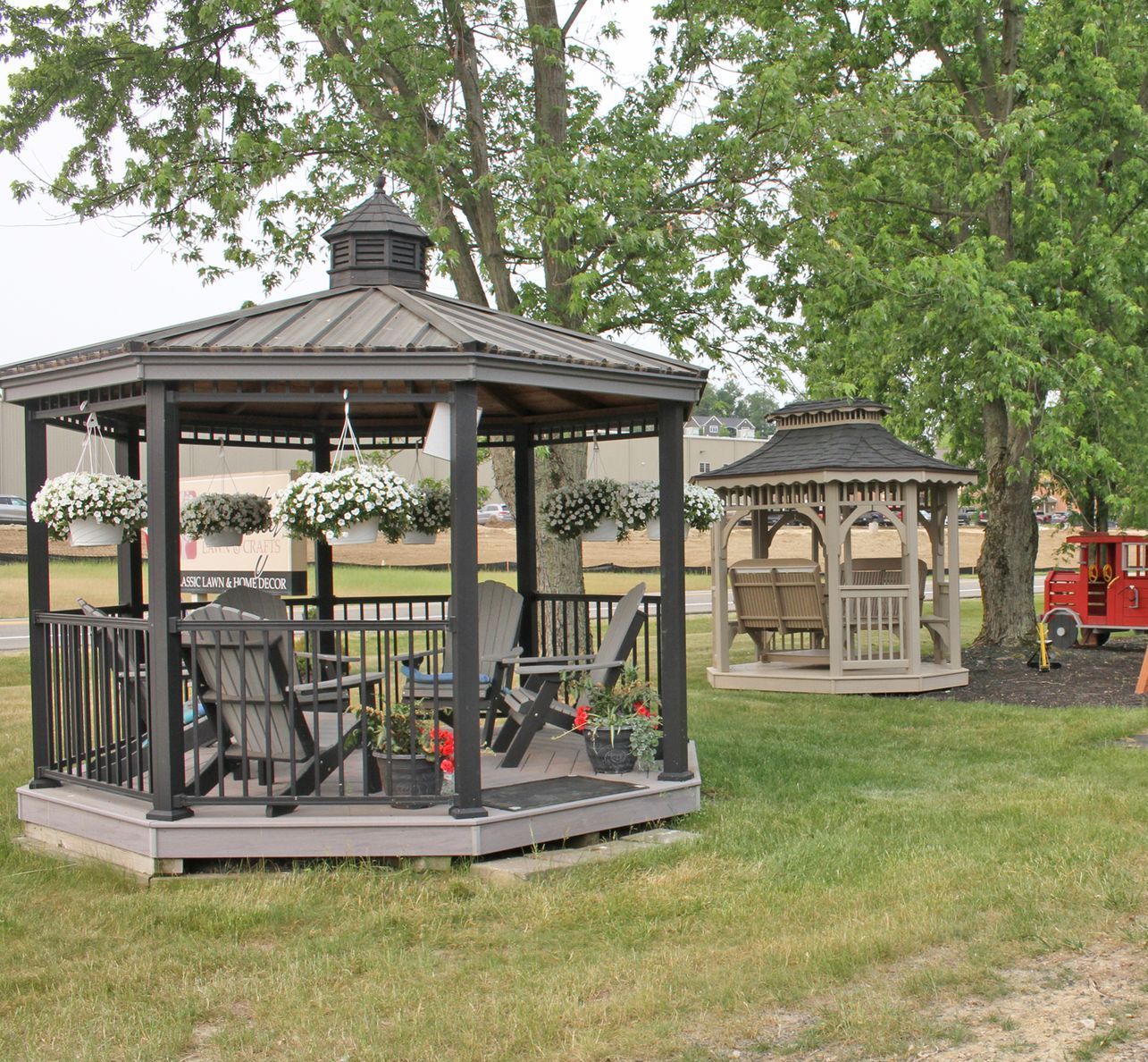 Two gazebos on grass, one black with chairs, the other tan with a swing. Trees and building in background.
