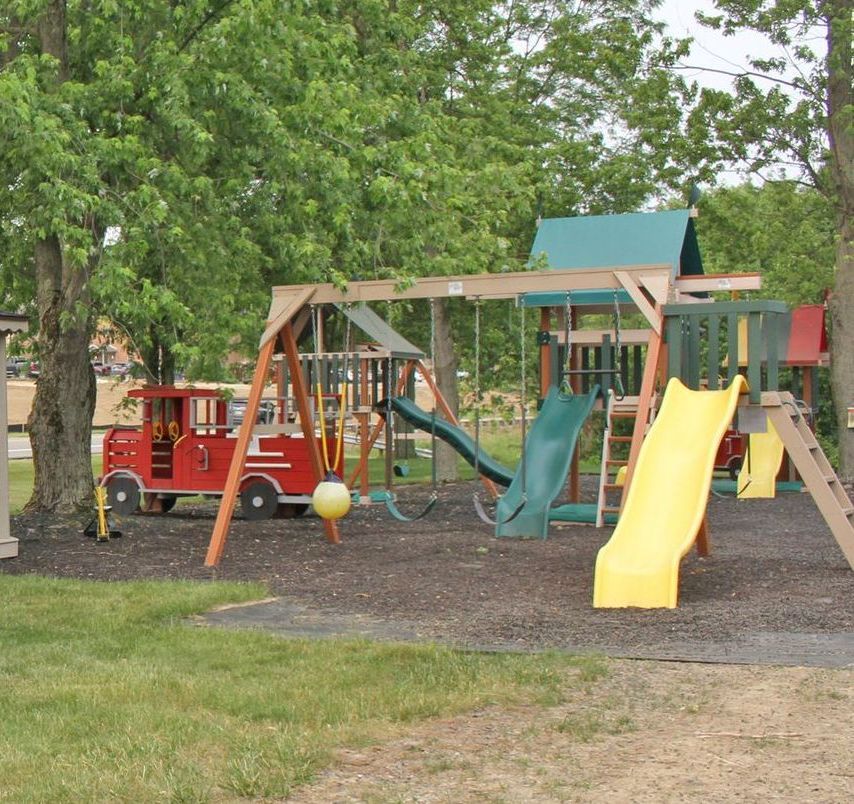 Playground with swings, slides, and a fire truck play structure on a bed of mulch, surrounded by grass and trees.