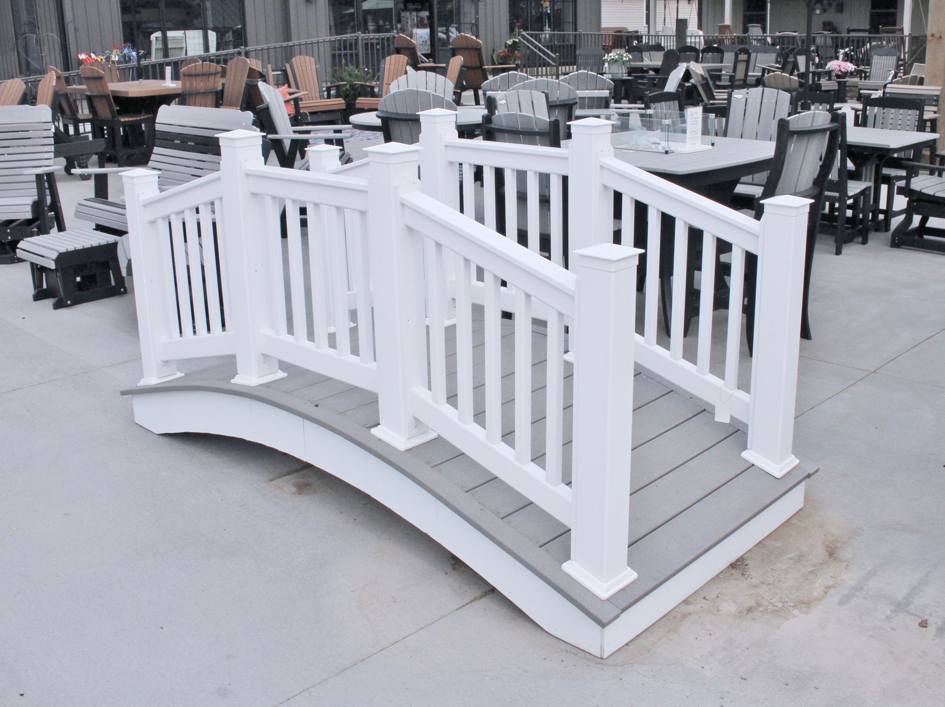 White arched bridge with gray decking and railings, surrounded by outdoor furniture.