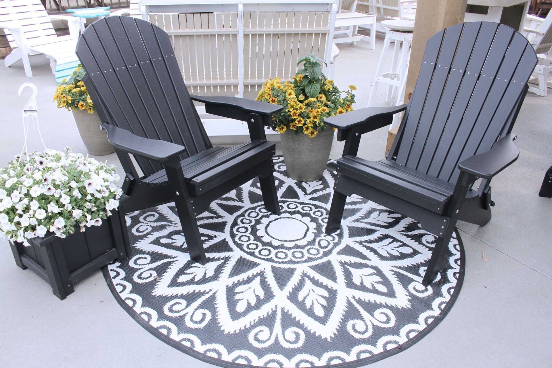Two black Adirondack chairs on a round patterned rug, potted flowers on either side.
