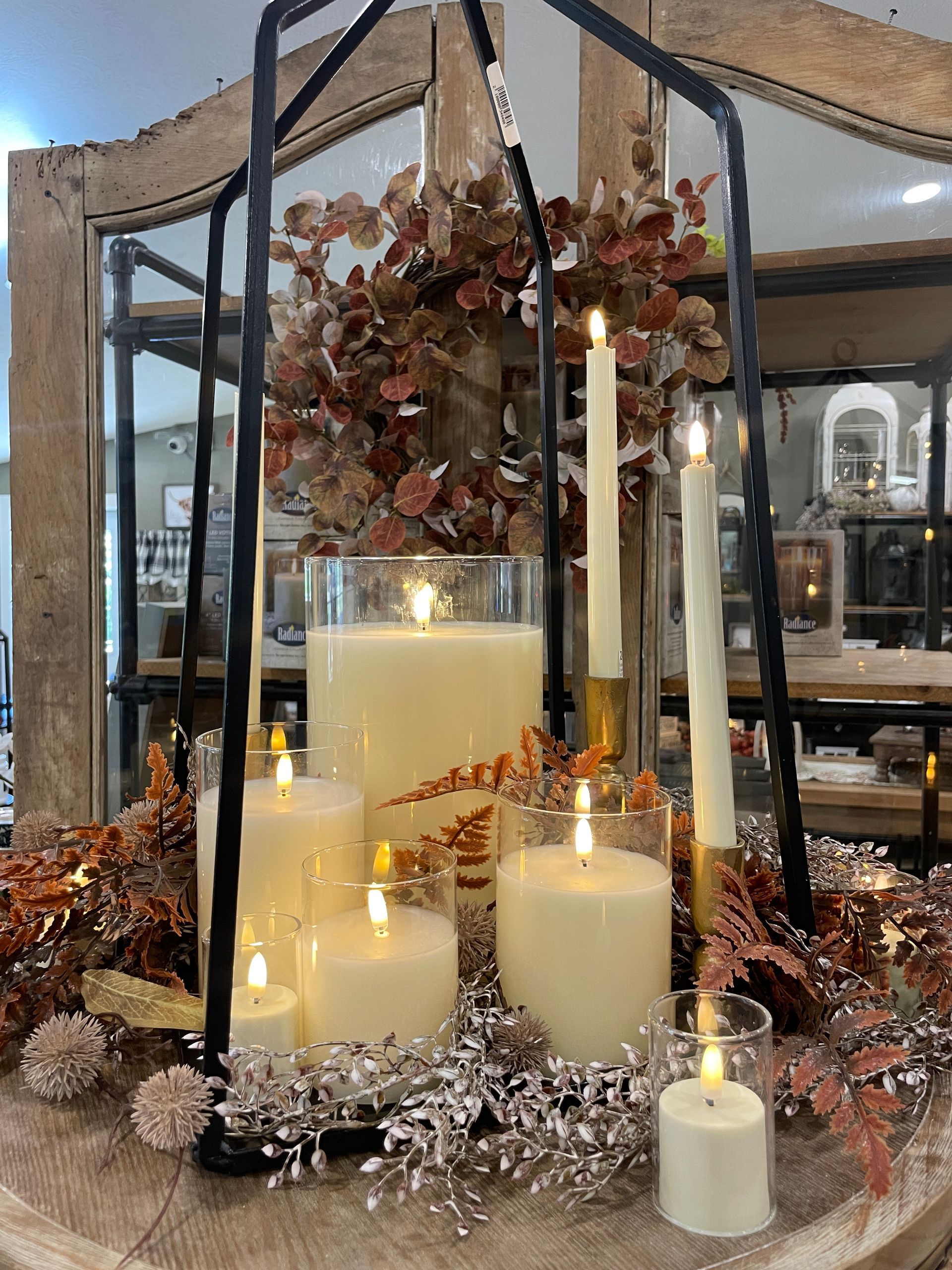 Arrangement of lit candles of various sizes inside a black lantern, with dried foliage and glass cylinders.