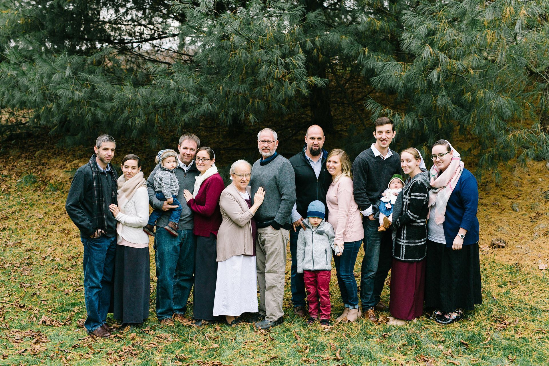 Family portrait outdoors, posing in front of leafy trees. People of various heights, wearing casual clothing, smiling.