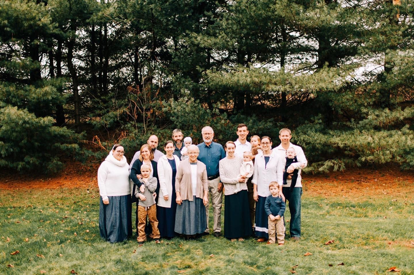 Group of people standing outdoors in front of trees. Some are holding babies, smiling.