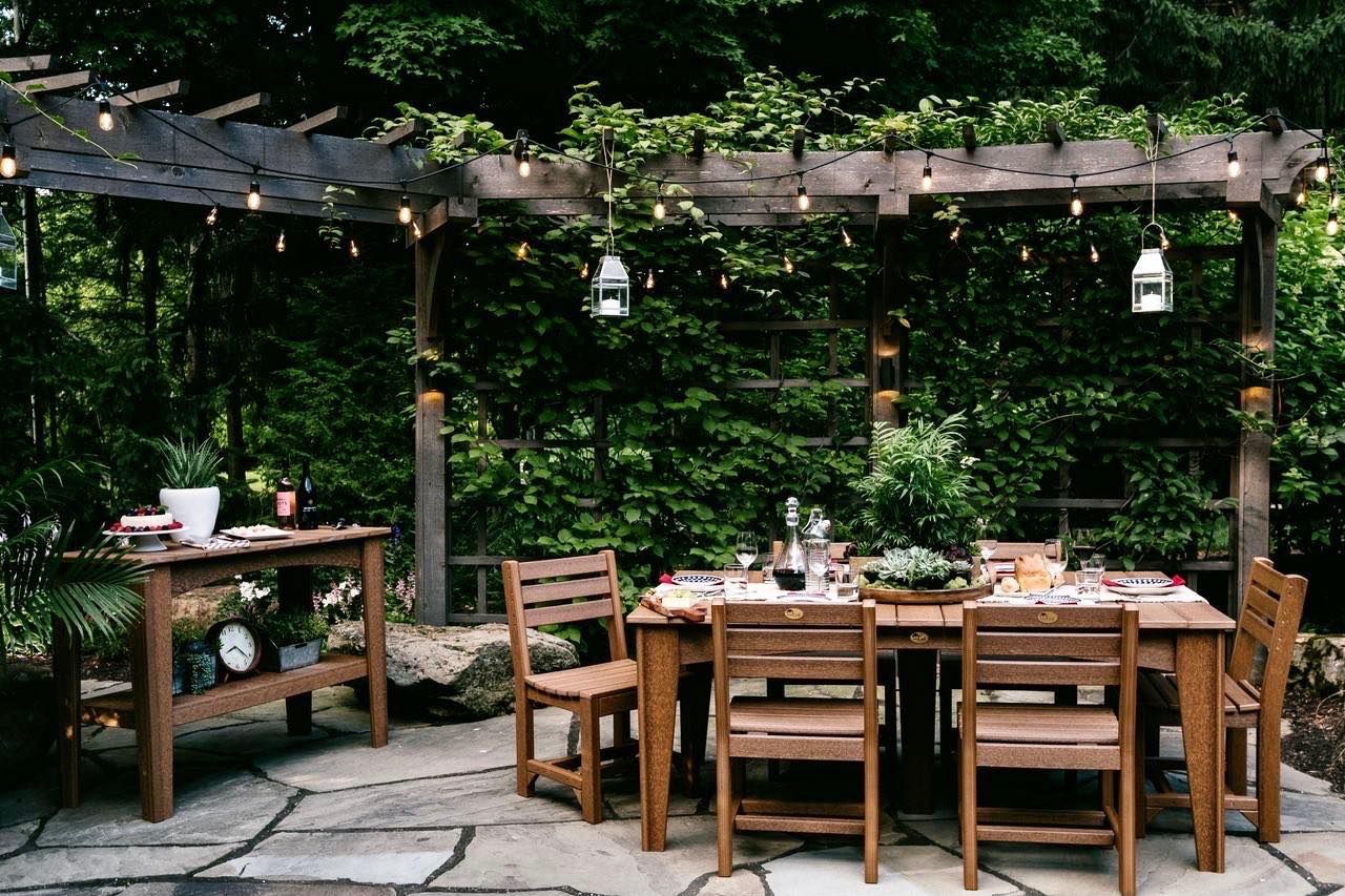 Outdoor dining table set beneath a wooden pergola strung with lights, surrounded by lush greenery.