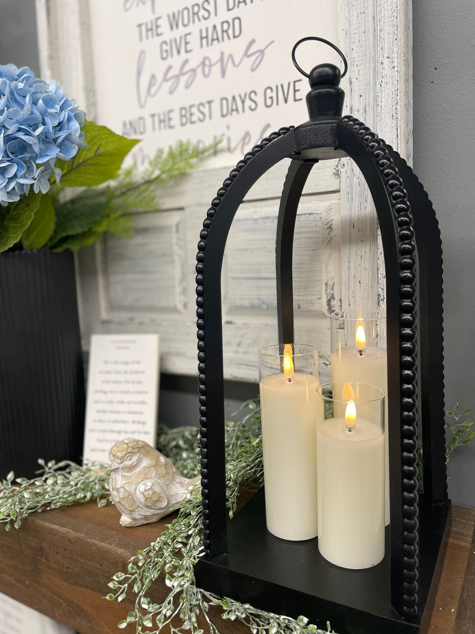 Black lantern with candles, flowers, and a sign on a shelf.