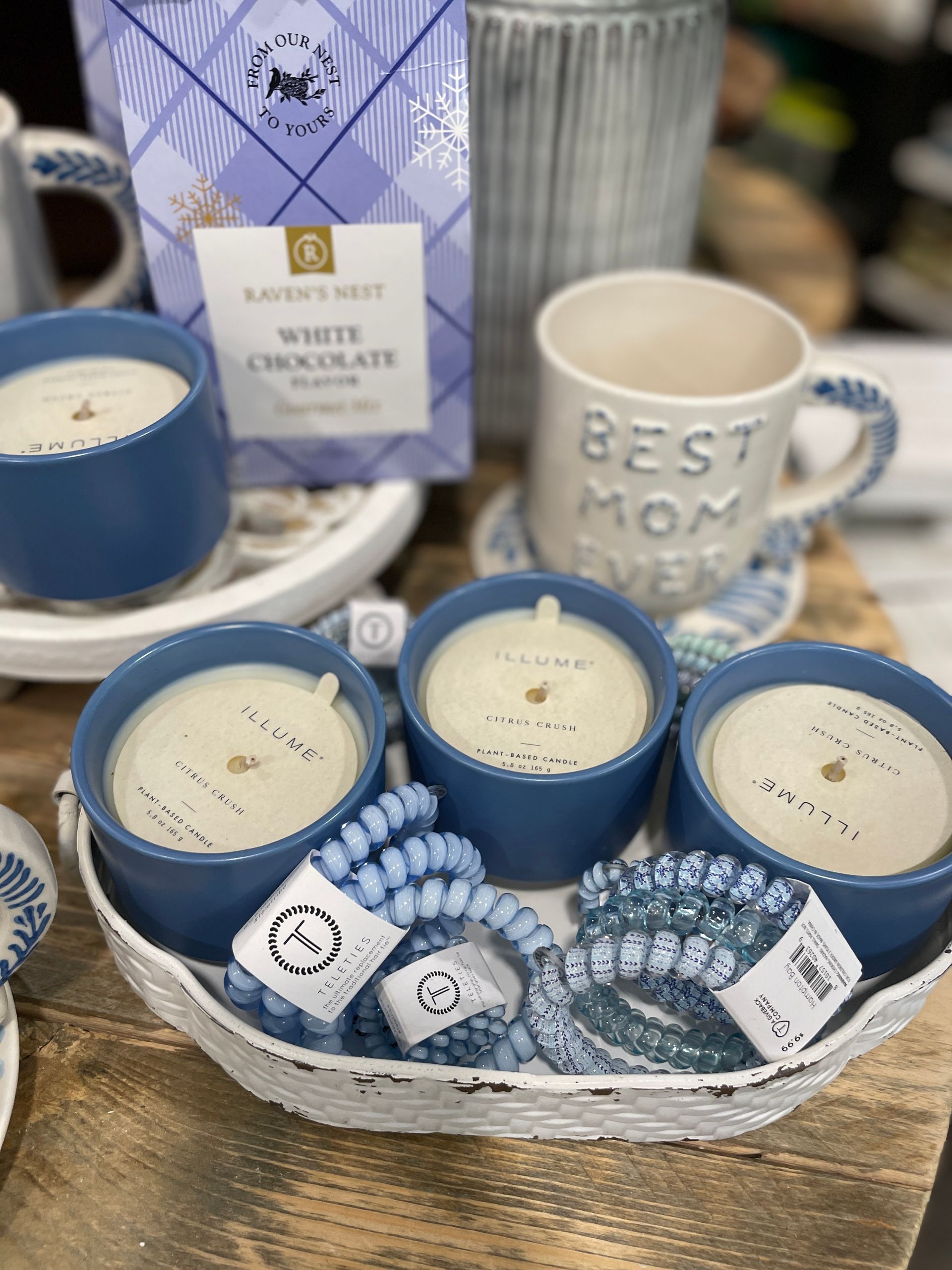 A white basket holds three blue candles, scrunchies, a mug, and hot chocolate.