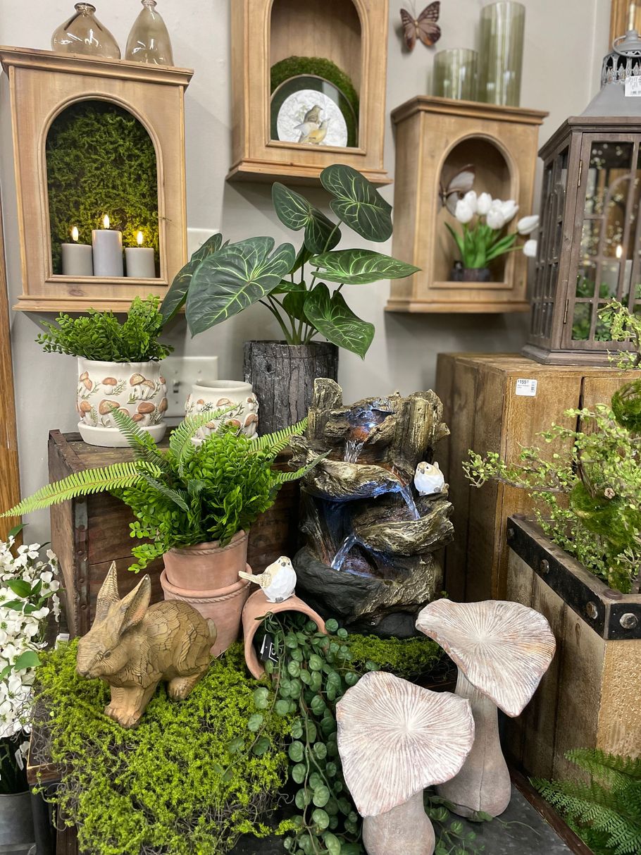 Display of potted plants, garden ornaments, candles, and a small fountain on shelves and rustic wood crates.