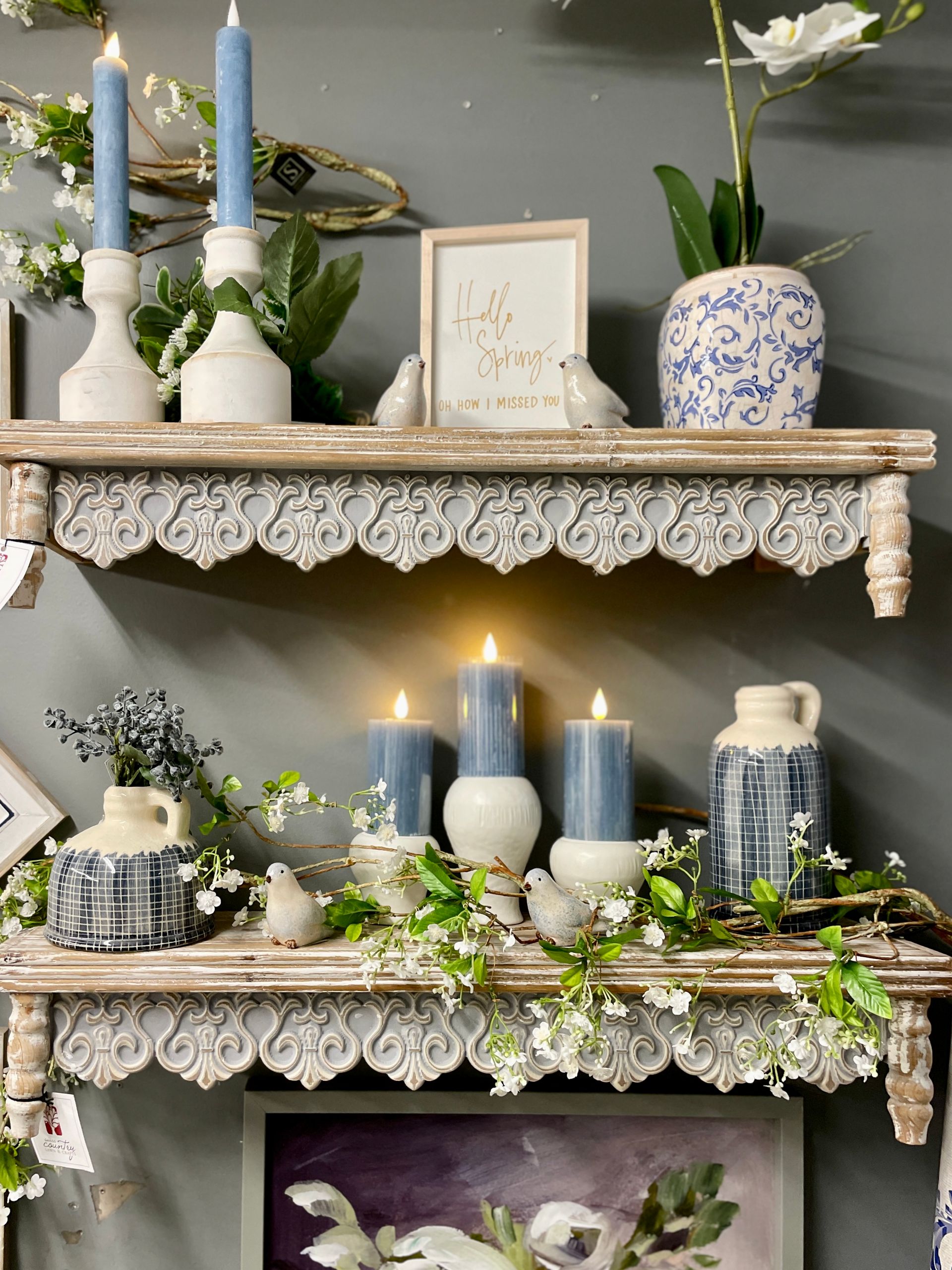 Two ornate, cream-colored shelves displaying candles, vases, and floral arrangements against a gray wall.