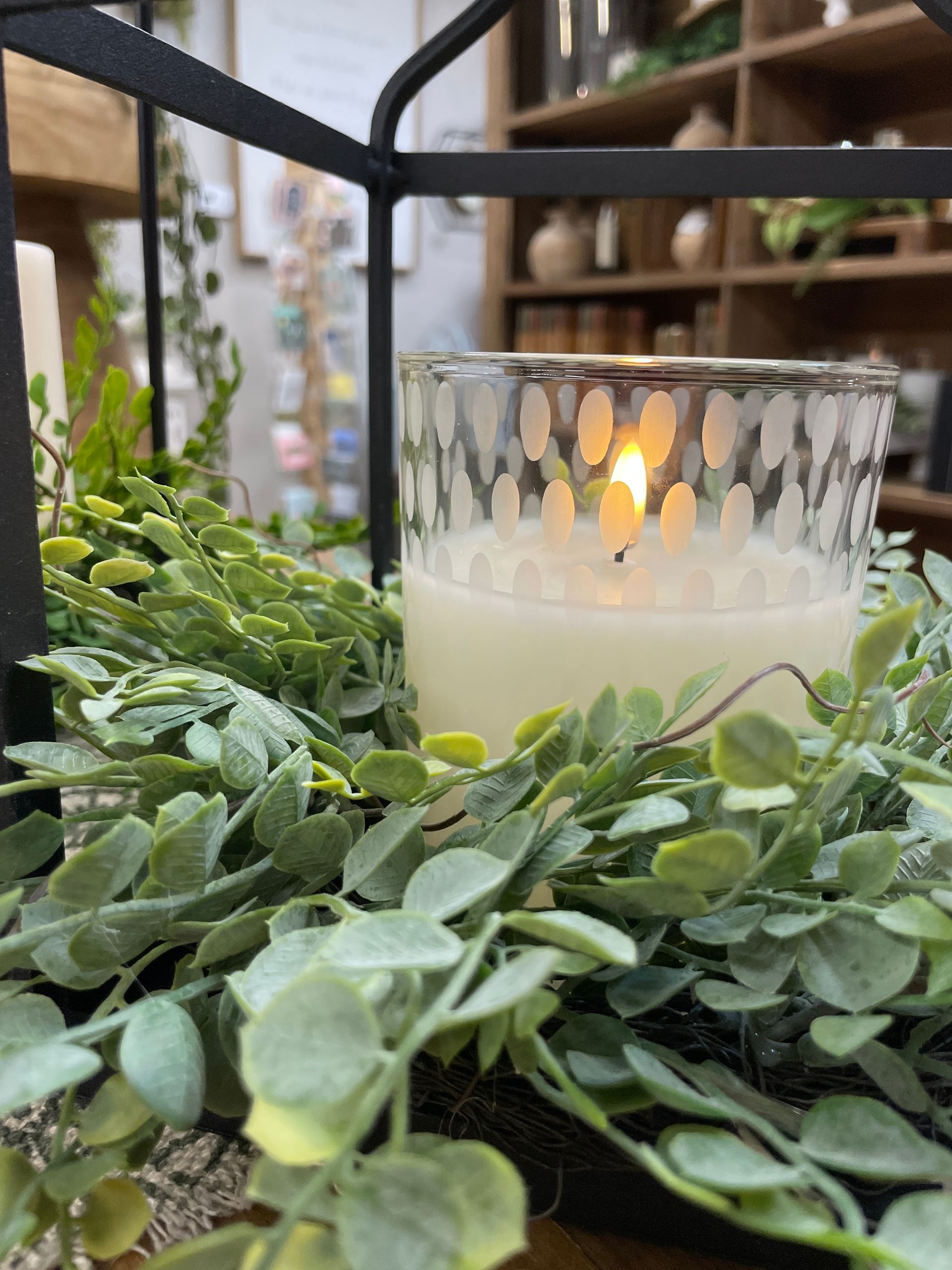 Candle lit inside a glass jar, surrounded by green foliage, inside a black metal frame.