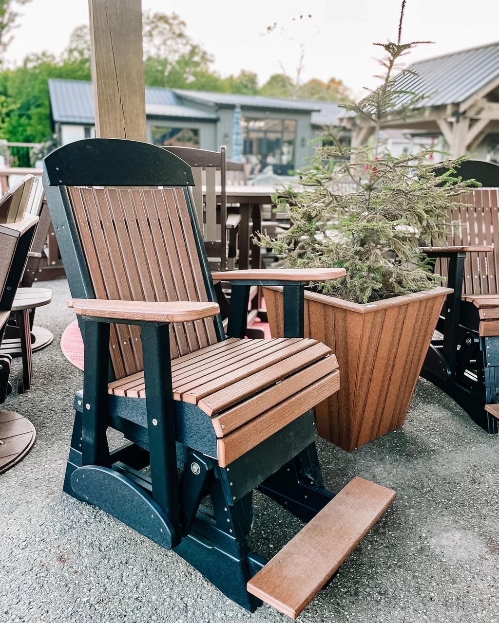 Black and brown outdoor rocking chair, with footrest, next to a brown planter filled with greenery.