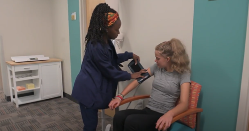 A medical professional adjusts a blood pressure cuff on a patient's upper arm while they sit in a clinical exam room.