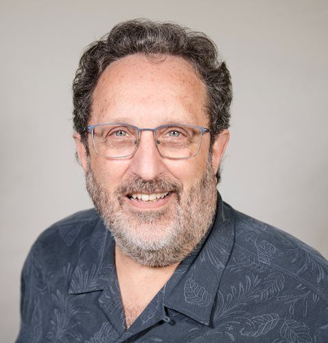 Headshot of a smiling person with curly brown hair, glasses, and a short beard, wearing a patterned dark blue shirt.